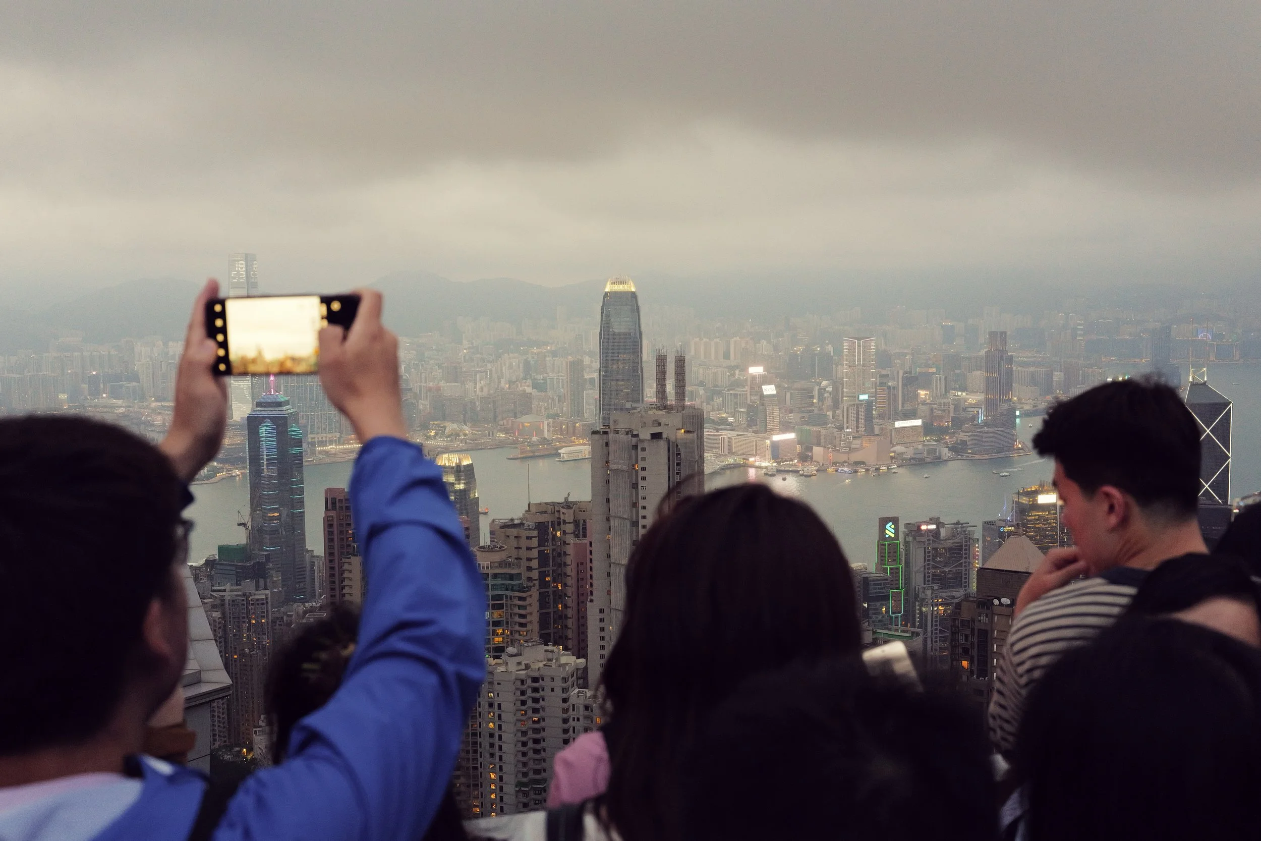 A group of people is gathered on a balcony or rooftop overlooking a city skyline, taking photos of the cityscape, which includes tall buildings, a river, and hills in the distance on a cloudy day.