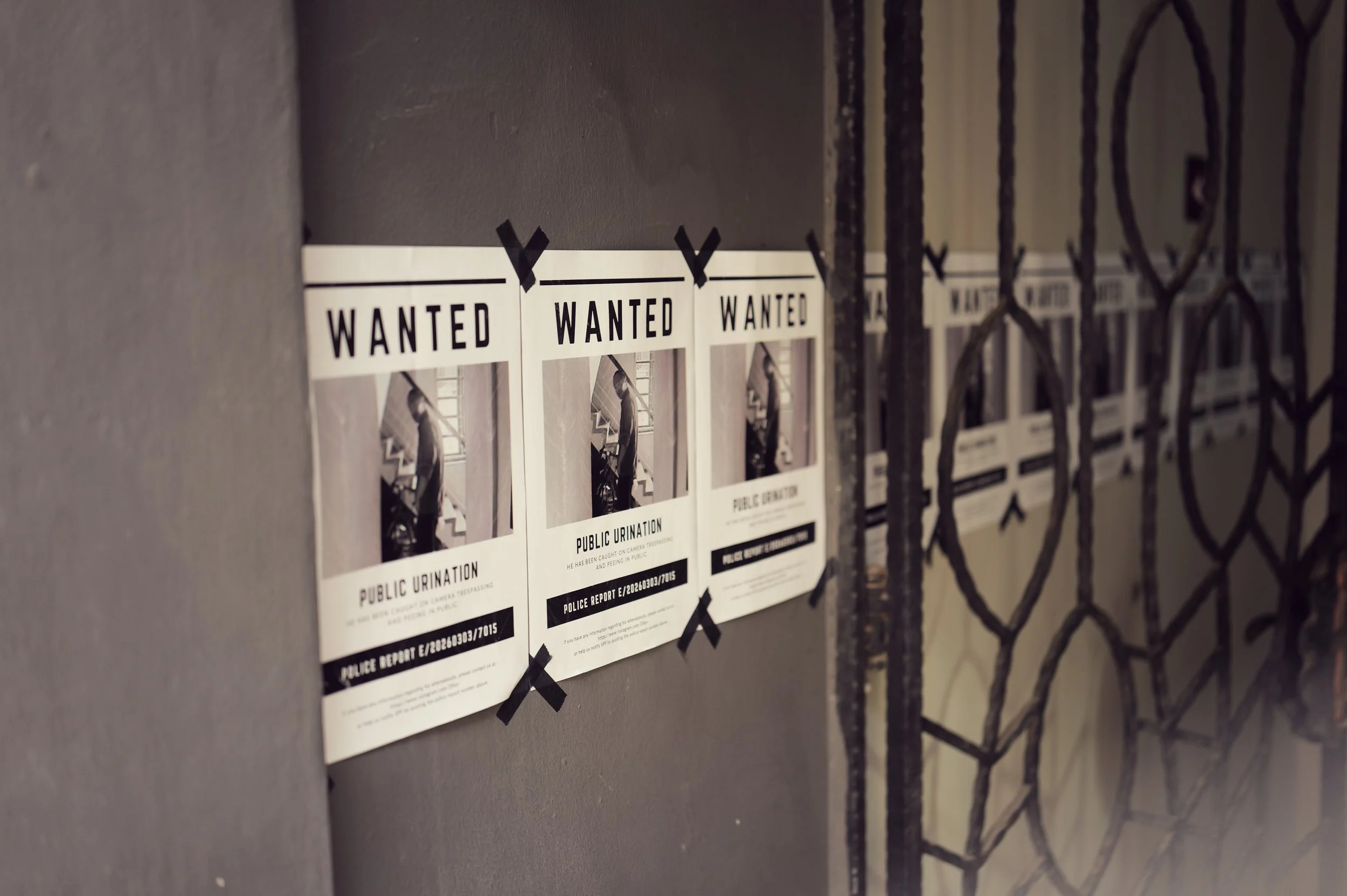 Several wanted posters on a wall behind a black metal gate. The posters feature a person and state 'Public Urination' along with a police report number.