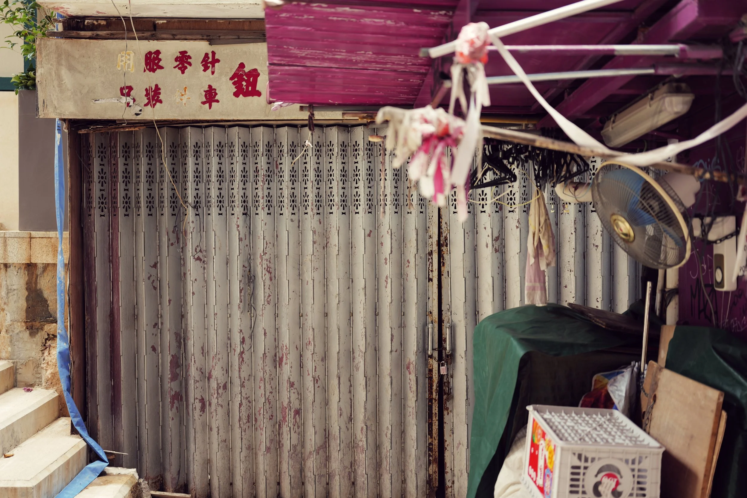 A cluttered outdoor space with old metal shutter, hanging clothes, fan, plastic crate, and various items, some with Chinese characters on a sign.