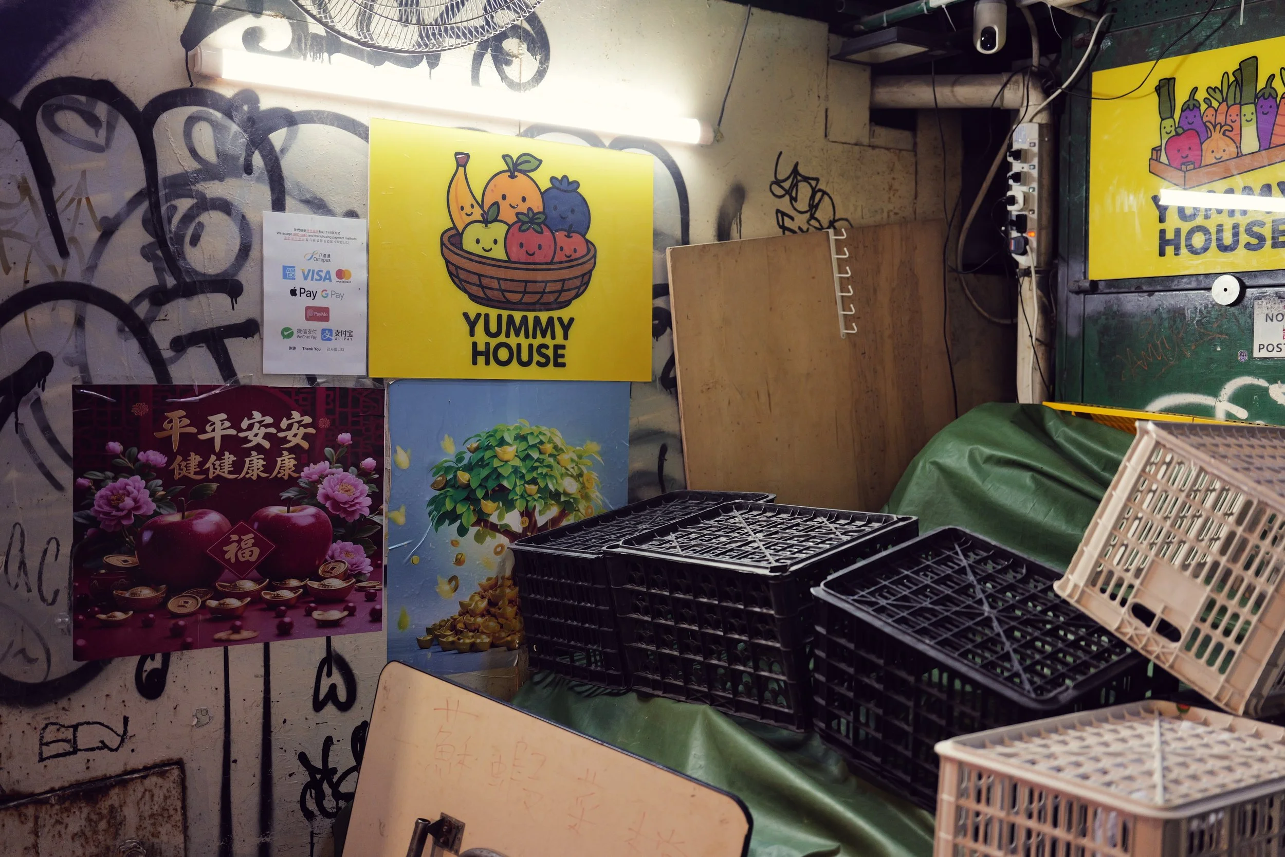 A yellow sign with a cartoon basket of smiling fruits including a banana, apple, blueberry, and strawberry with the text 'YUMMY HOUSE' in black. There are various other signs, some with flowers and Chinese characters. Black plastic crates and white p