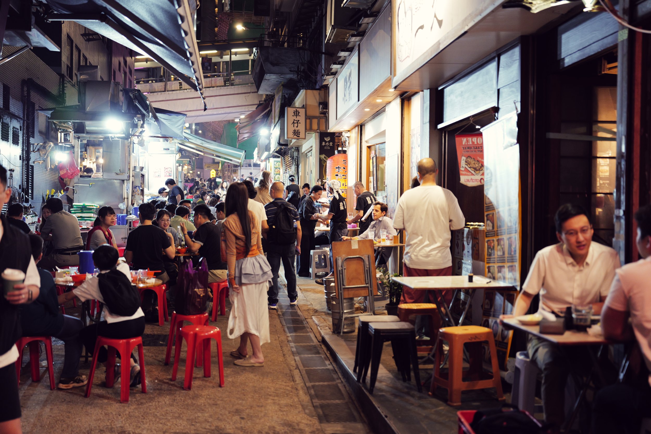 A bustling outdoor night market with many people sitting at tables and ordering food from vendors. The scene is lively with some standing and others dining, illuminated by warm lighting.