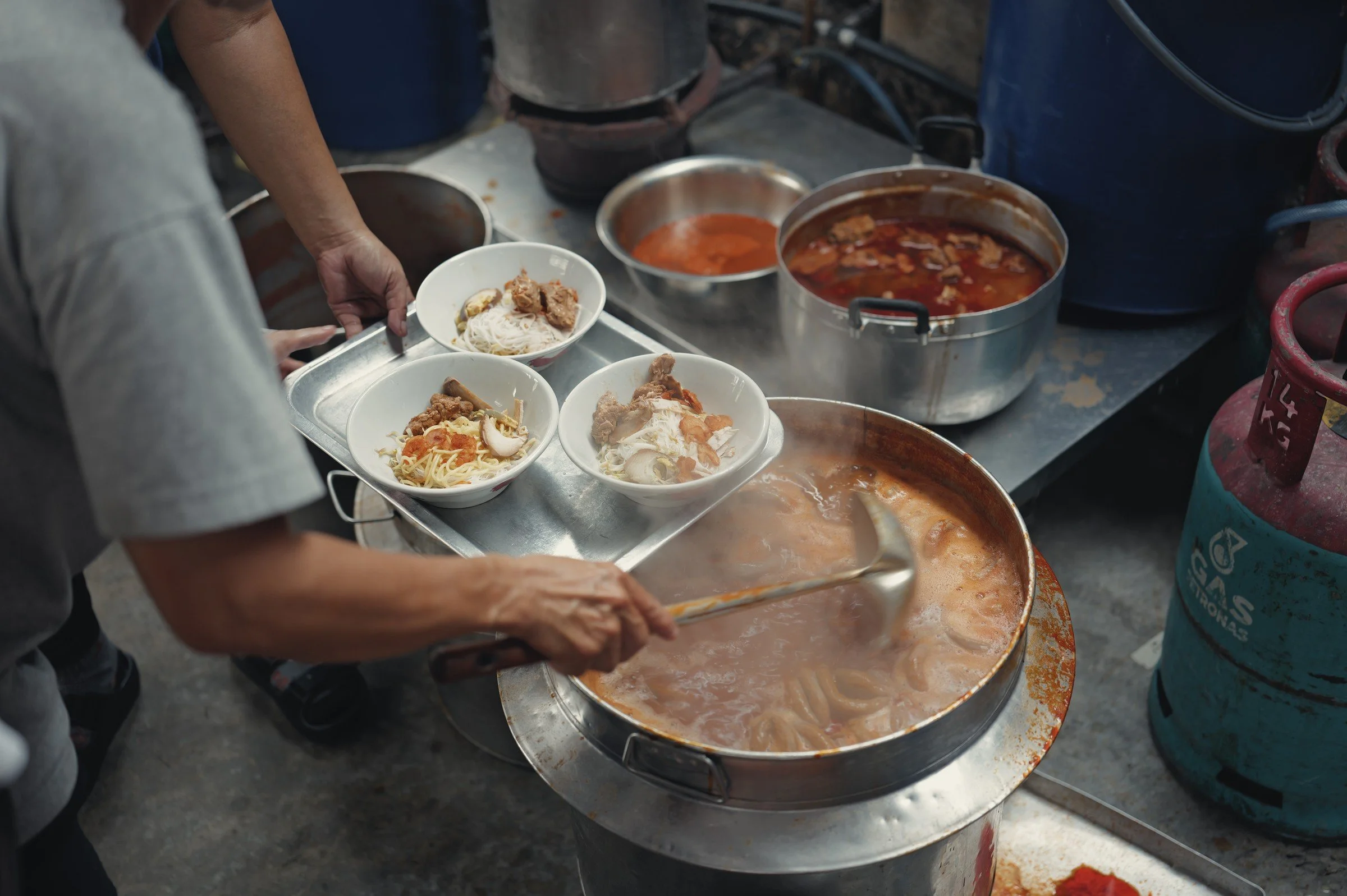 A cook serves bowls beside a wide steaming pot at a food stall.