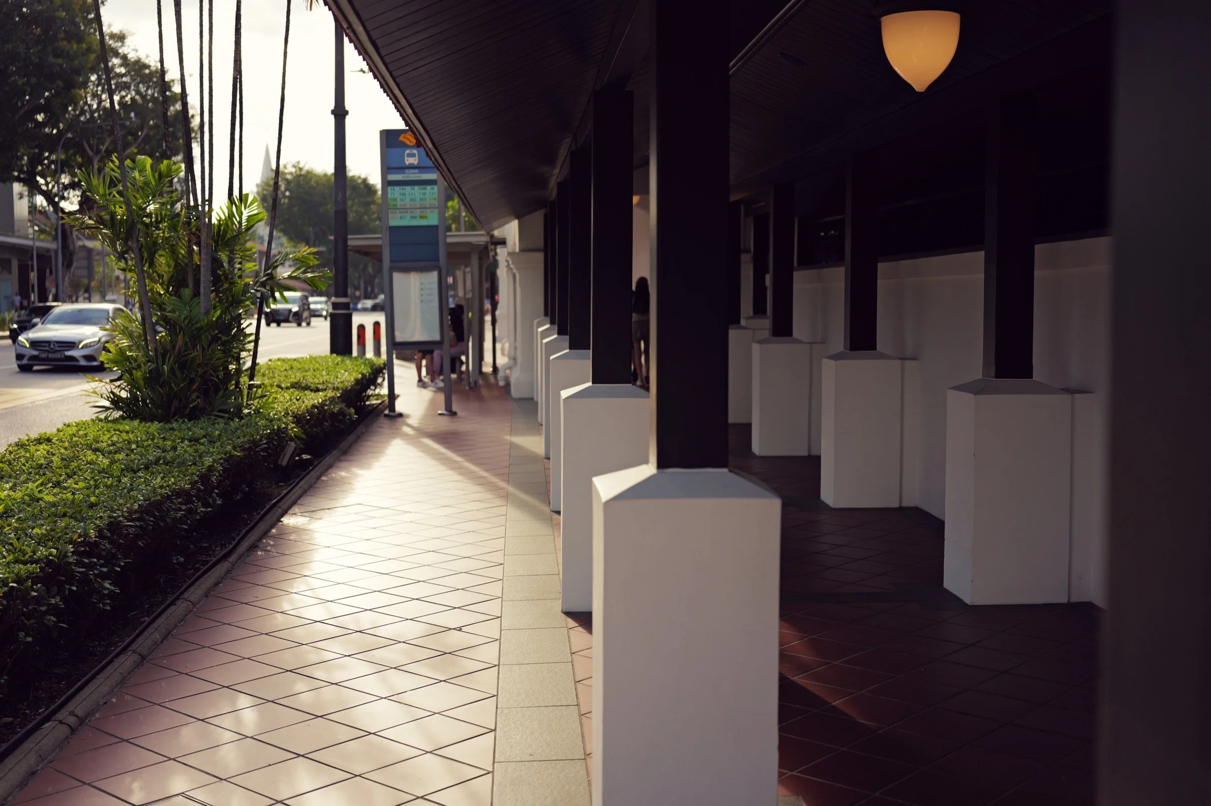 A shaded walkway with white columns and red floor tiles opens onto roadside greenery and palms.