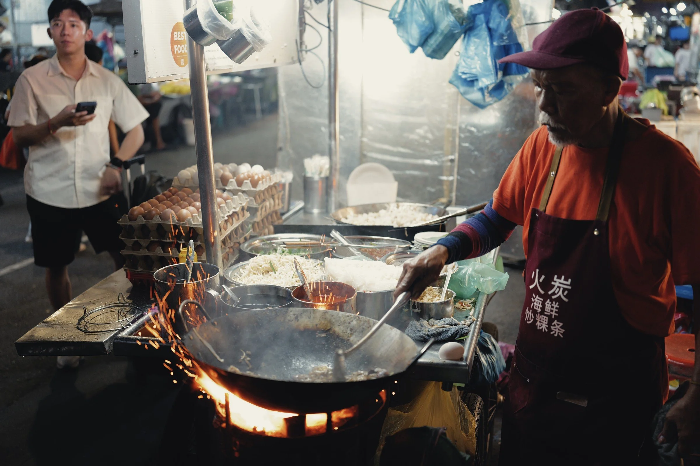 Sparks rise beside a hot wok as a street-food cook works over an open flame.