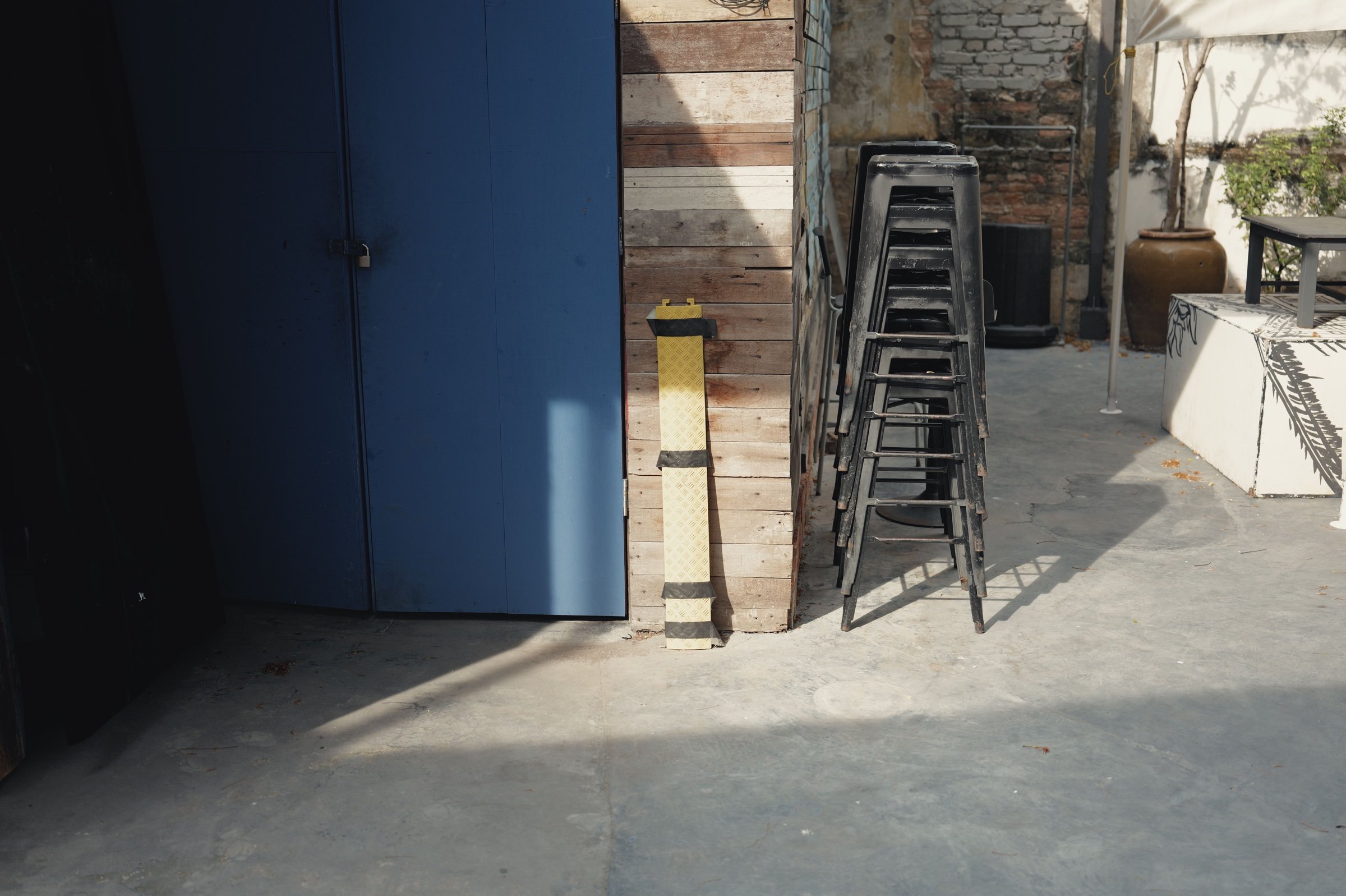 Stacked stools stand beside a blue door in slanting sunlight.