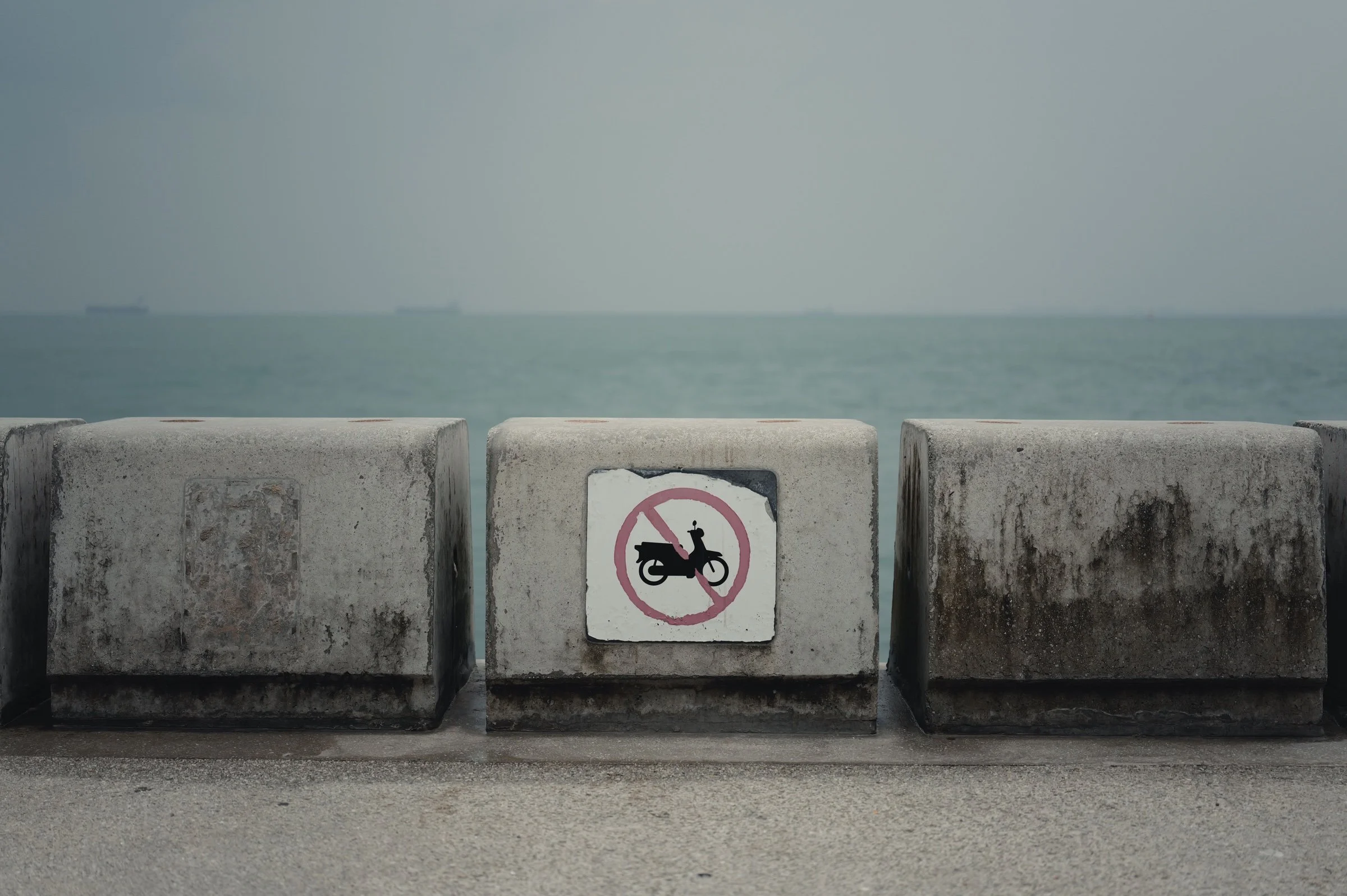 A faded no-motorcycle sign marks a weathered seawall beside a hazy sea.