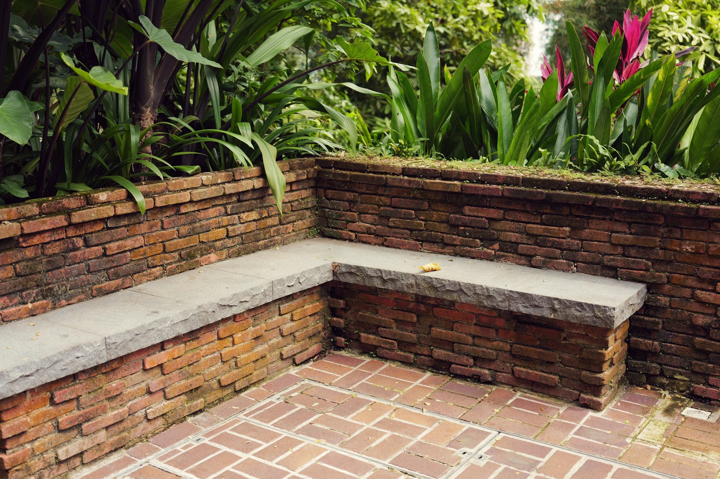 An L-shaped brick and stone bench sits beneath tropical foliage at Fort Canning Park, Singapore.
