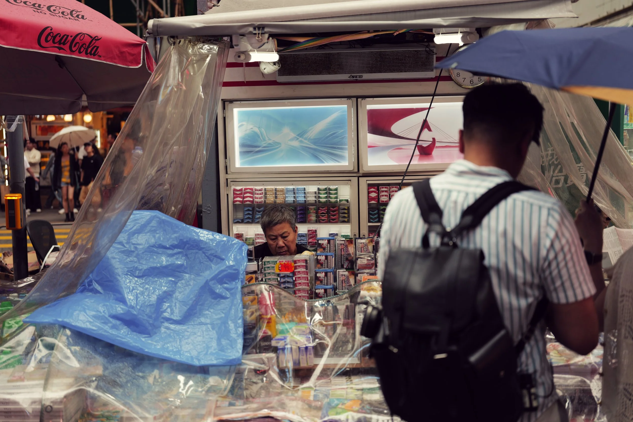 A street vending stand selling various small items, with a man and a woman customer in front, and people with umbrellas in the background.