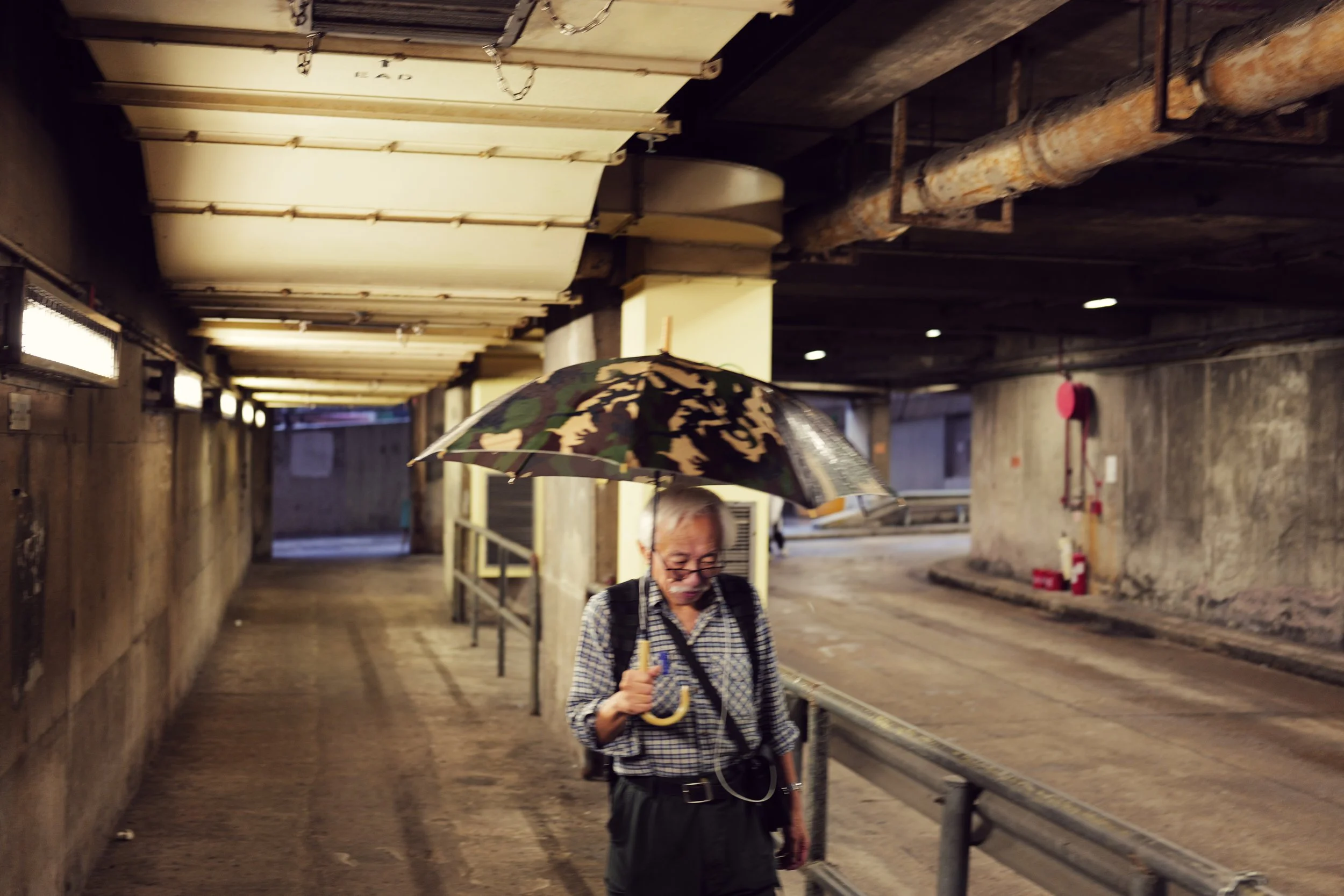 An elderly woman with glasses and a plaid shirt is holding an umbrella and walking in a covered outdoor corridor, with industrial pipes overhead and concrete walls on either side.