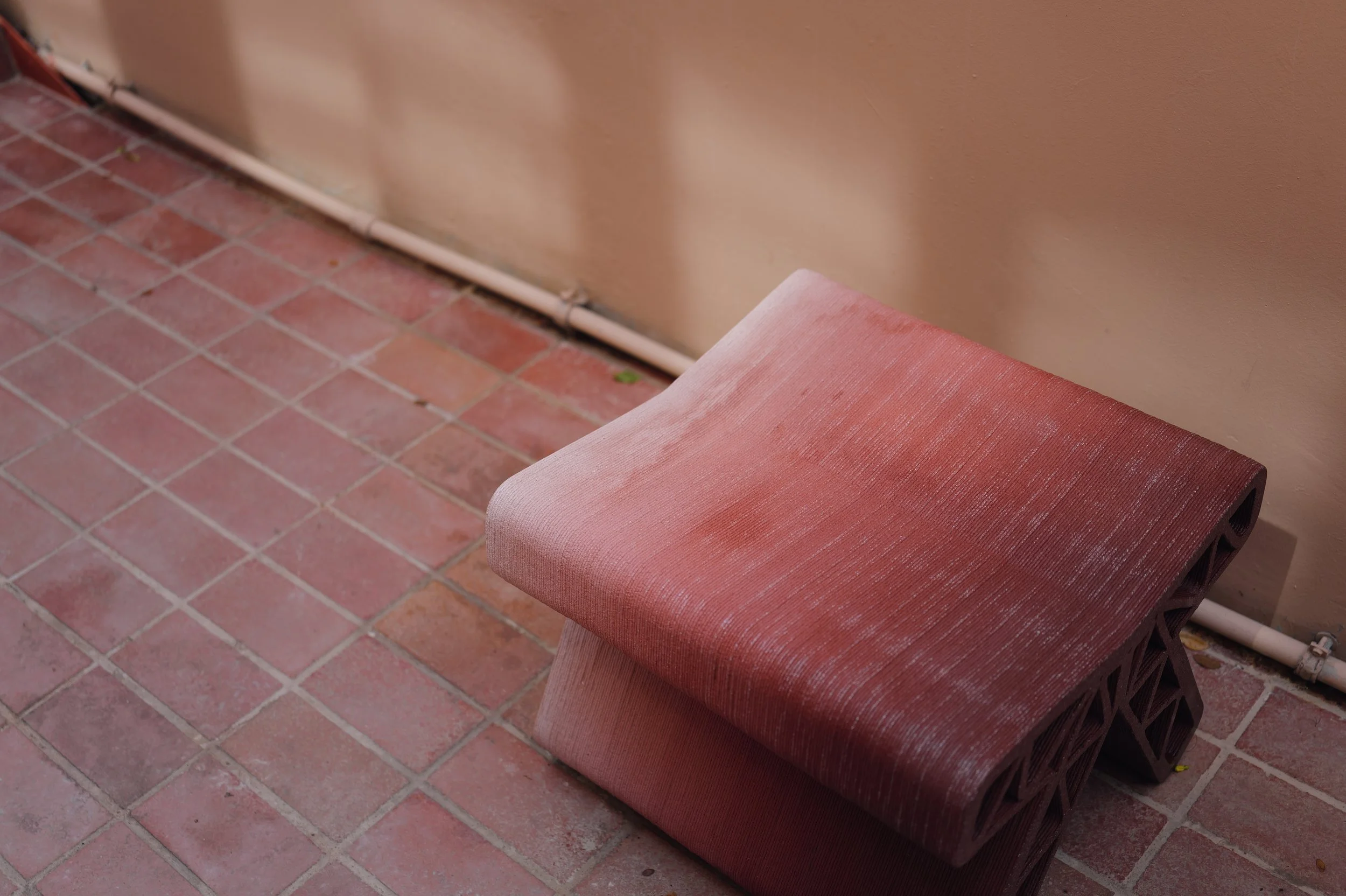 A pink cushioned bench with a textured fabric sits against a beige wall with a baseboard heater and a pipe. The floor is made of reddish-brown tiles with visible grout lines.