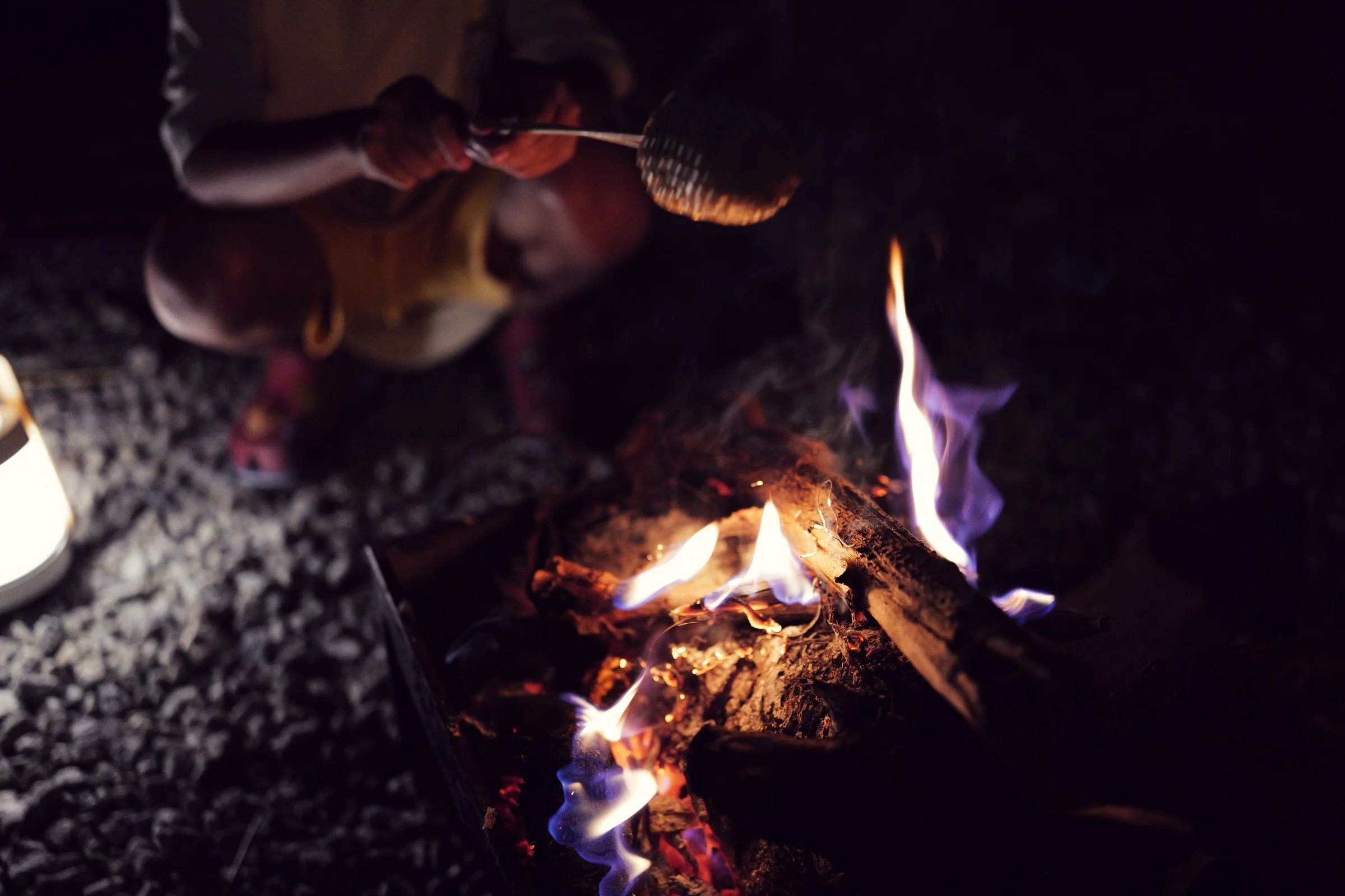 Hands hold food over a small campfire beside a glowing lantern.