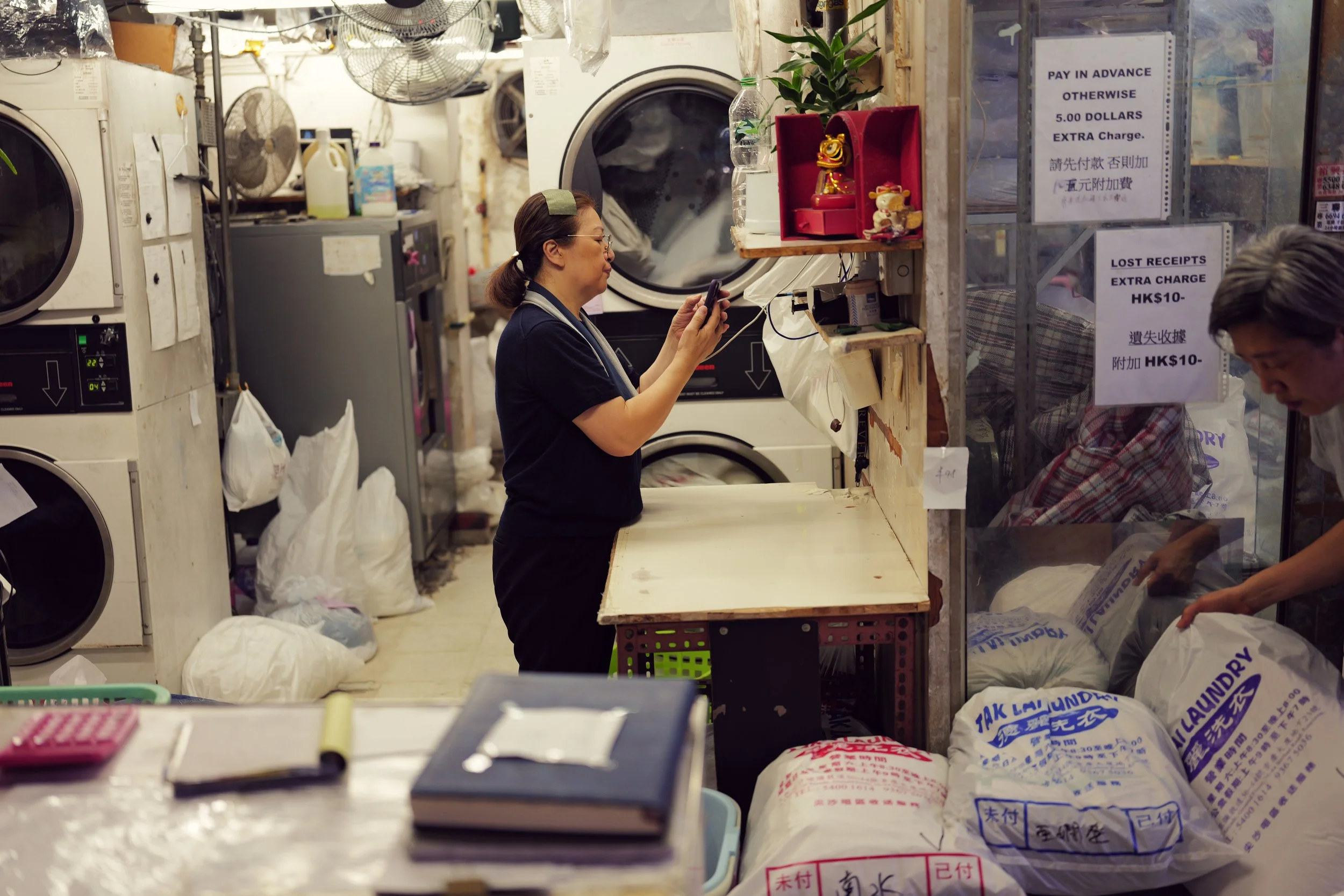 A laundromat with a woman in a black shirt using her phone while standing at a counter. Behind her are washing machines, dryers, and various laundry supplies, with bags of laundry on the floor. There are also signs with payment and fee information on