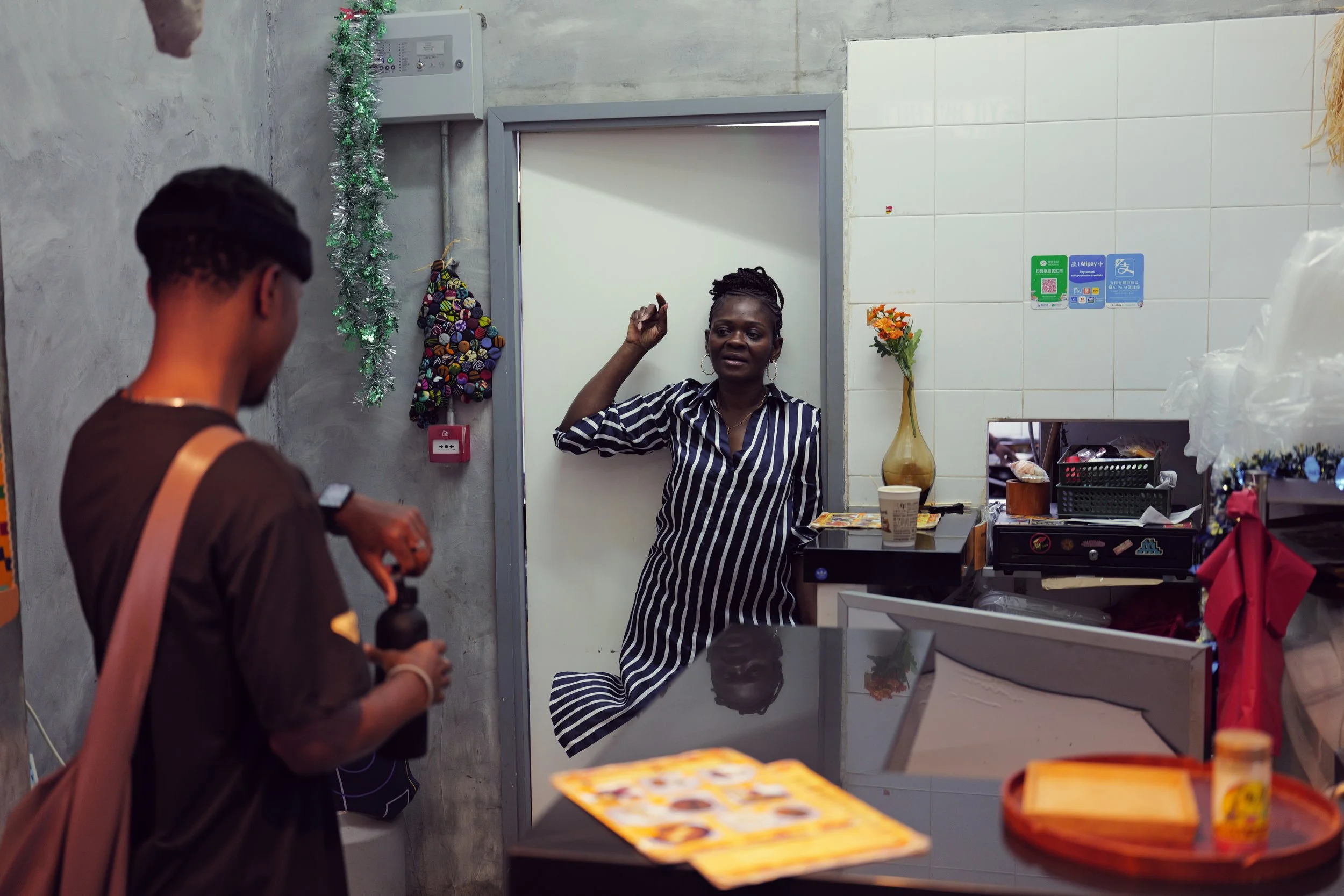 A woman with braided hair wearing earrings and a striped shirt is standing behind a counter, having a conversation with a man holding a camera, in a small shop decorated with tinsel, a colorful crocheted bag, and a vase with orange flowers.
