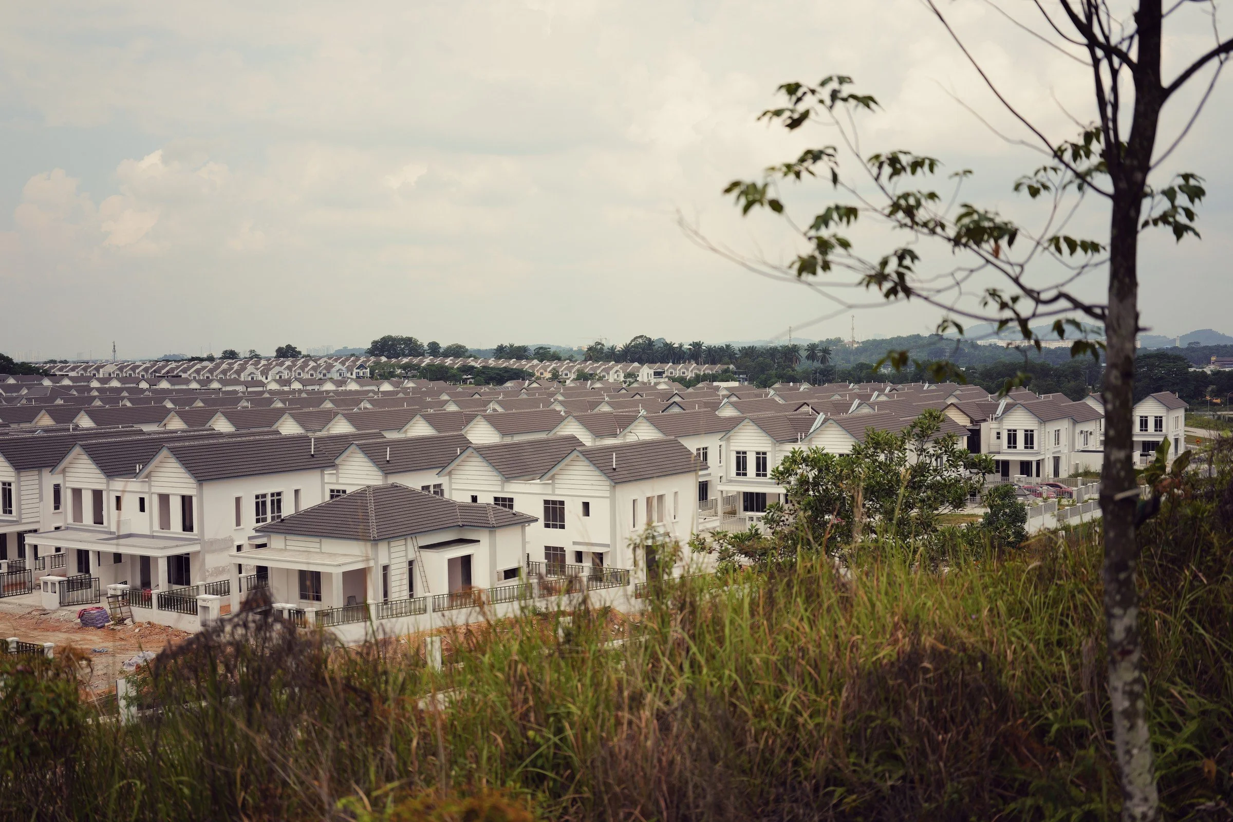 Rows of white houses and grey roofs repeat beneath a cloudy sky, seen through hillside trees.