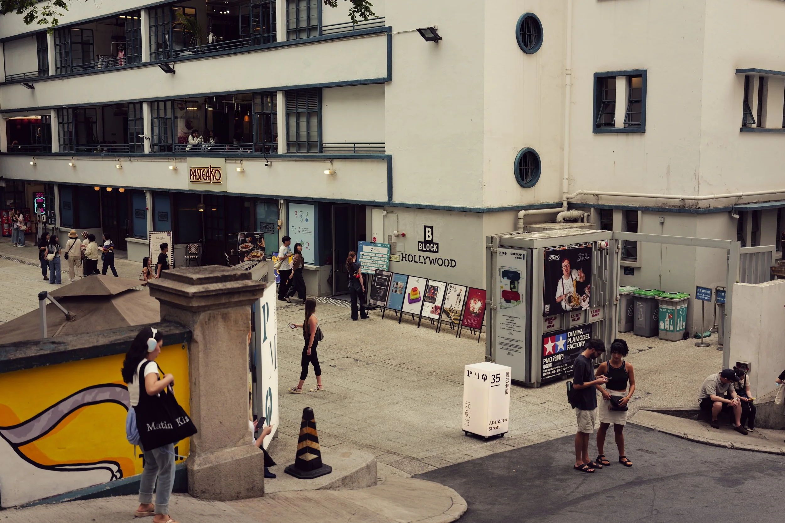 A busy street scene at Aberdeen Street in Hollywood, showing pedestrians walking, some standing and chatting, near storefronts and signage, including a 'Pastakao' restaurant, and posters advertising local businesses, with an apartment building above 