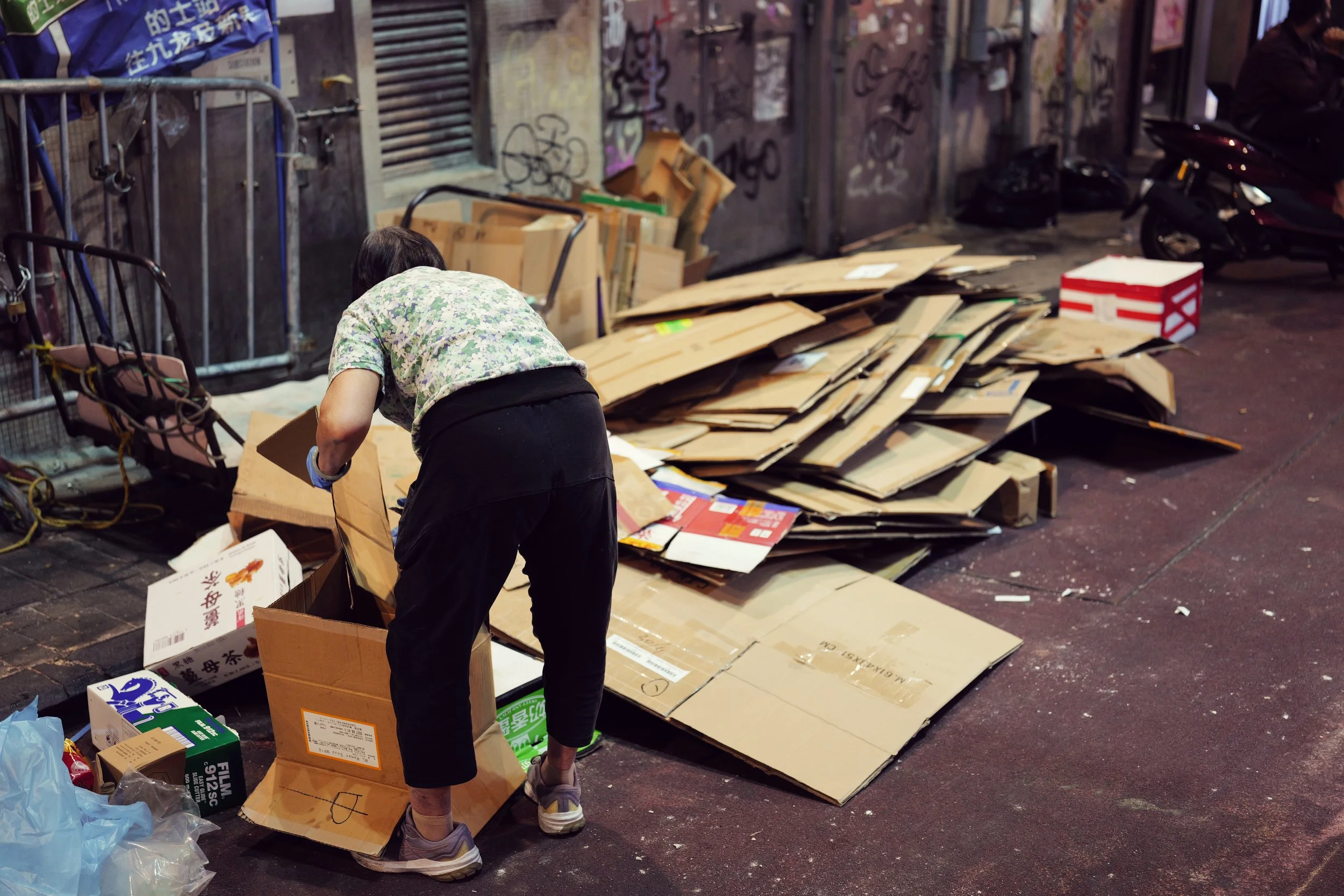 A person kneeling on the ground surrounded by cardboard boxes and paper, on a city sidewalk with graffiti-covered walls and parked motorcycles.