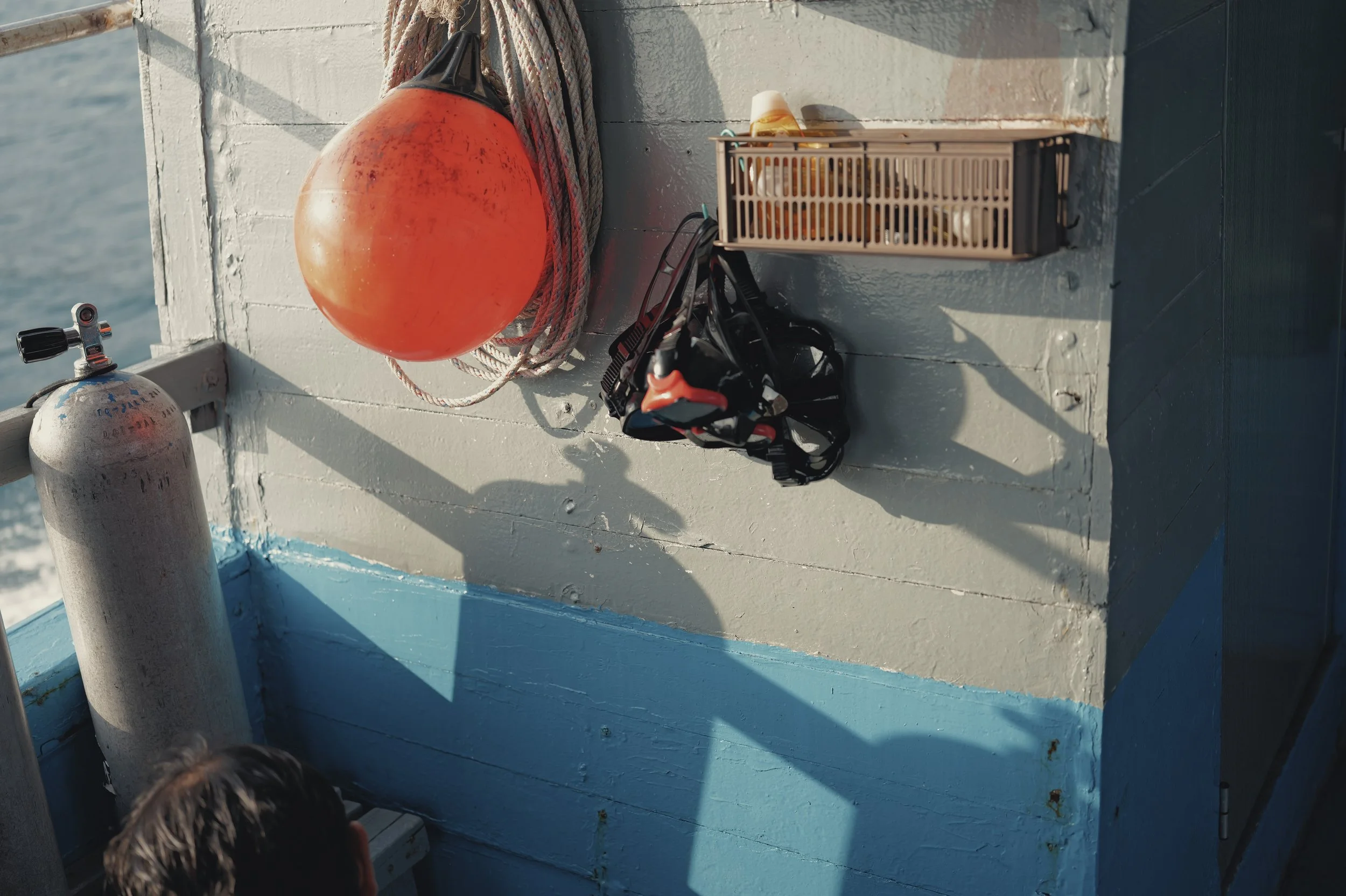 View of the side of a boat showing a large orange buoy, a rope, a beige storage container with straps, a black harness or strap, and a person's head with dark hair.