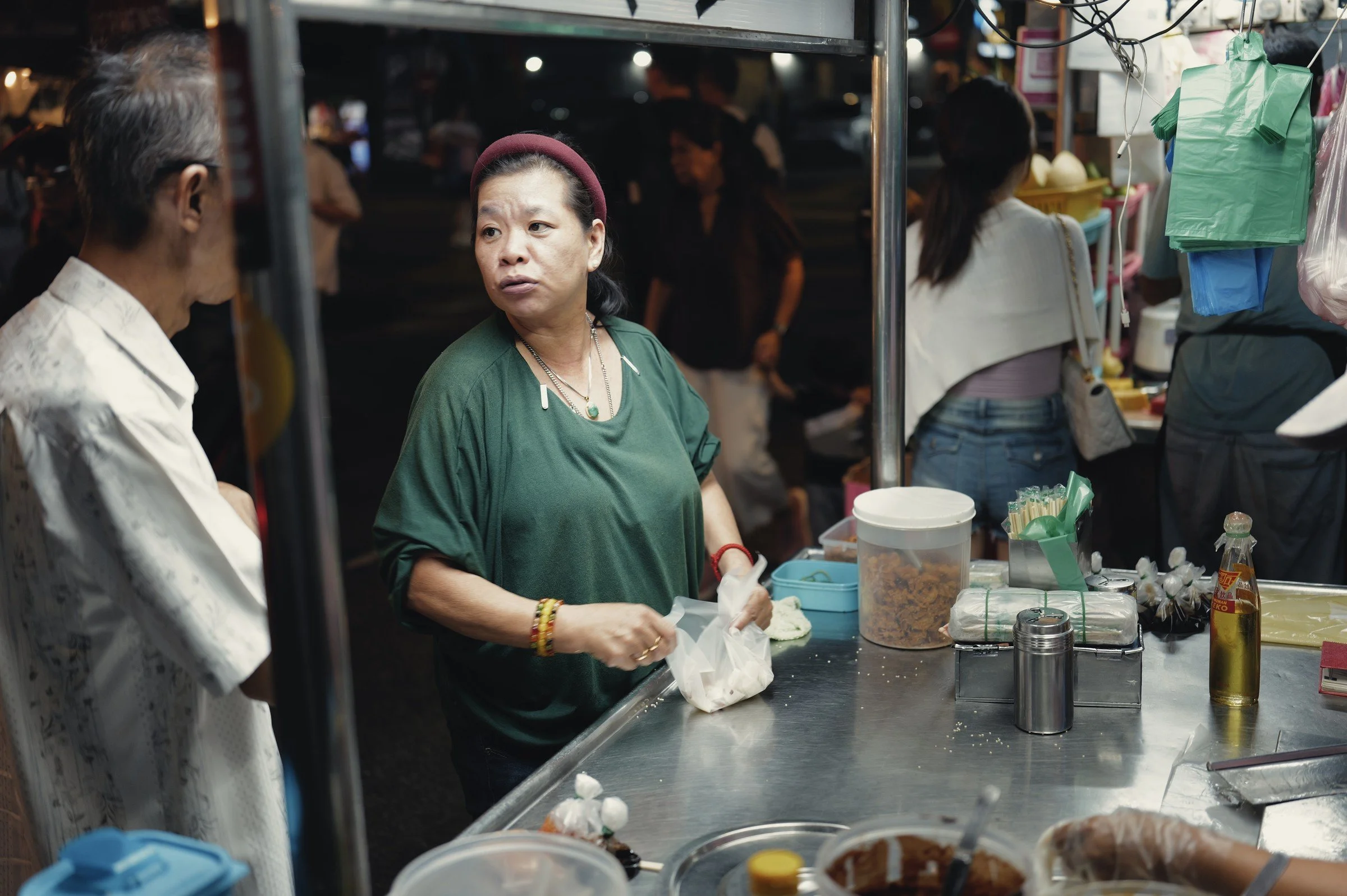 Two people talk across a metal food-stall counter surrounded by cups and containers.