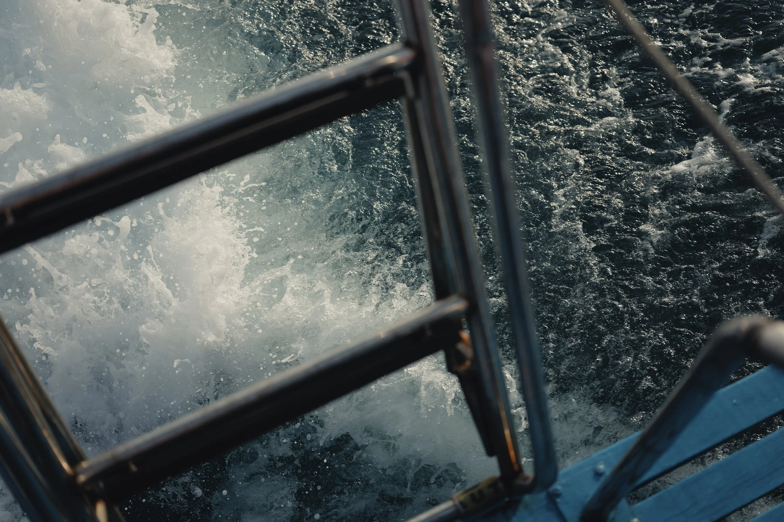 View of water crashing against a boat's metal railing, with the boat moving through the water, creating white foam and waves.