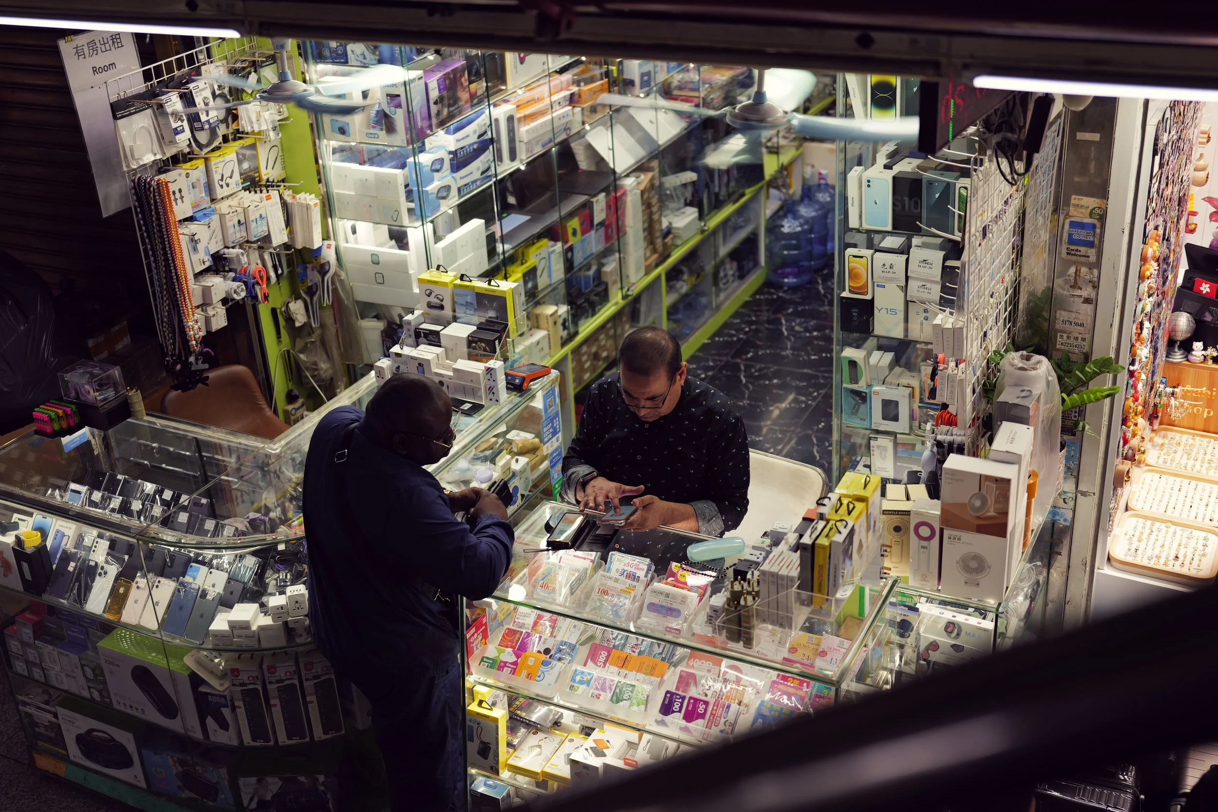 View of a store selling electronics and accessories, with two men at the counter examining products inside. The store shelves are filled with boxes of gadgets, electronic devices, and accessories in clear display cases. The photo is taken through a w