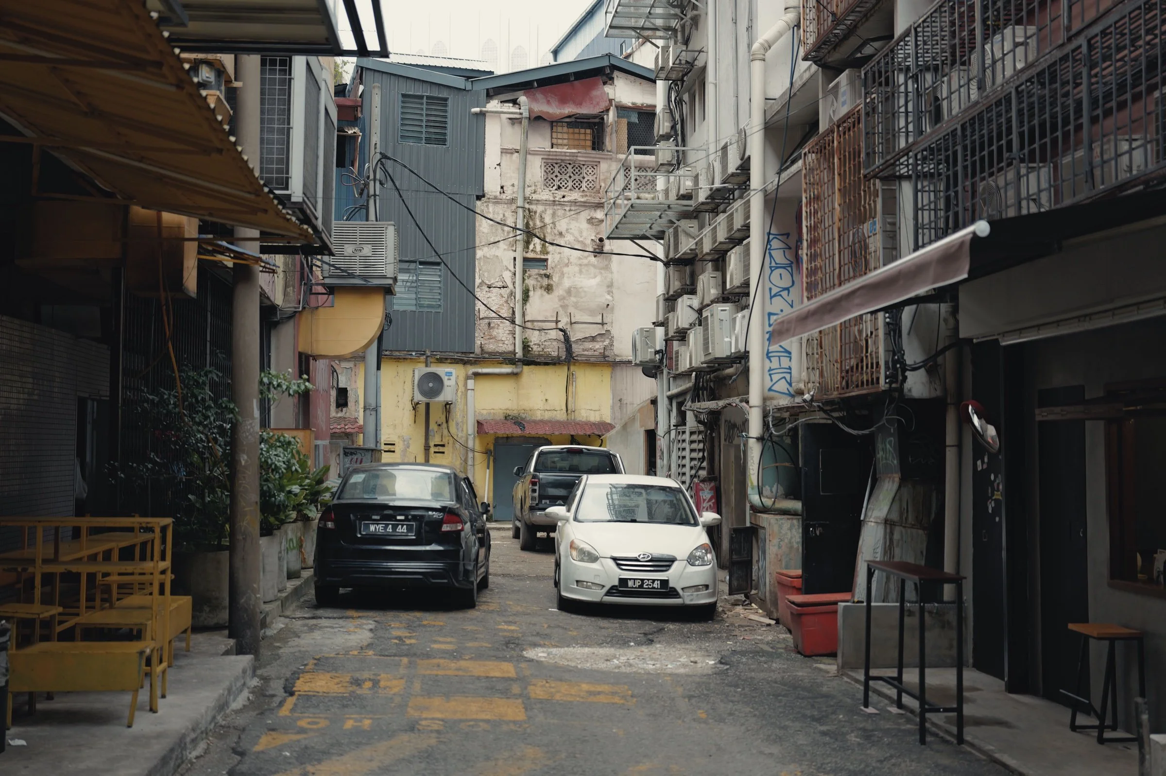 Parked cars line a narrow alley beneath wires and air-conditioning units.