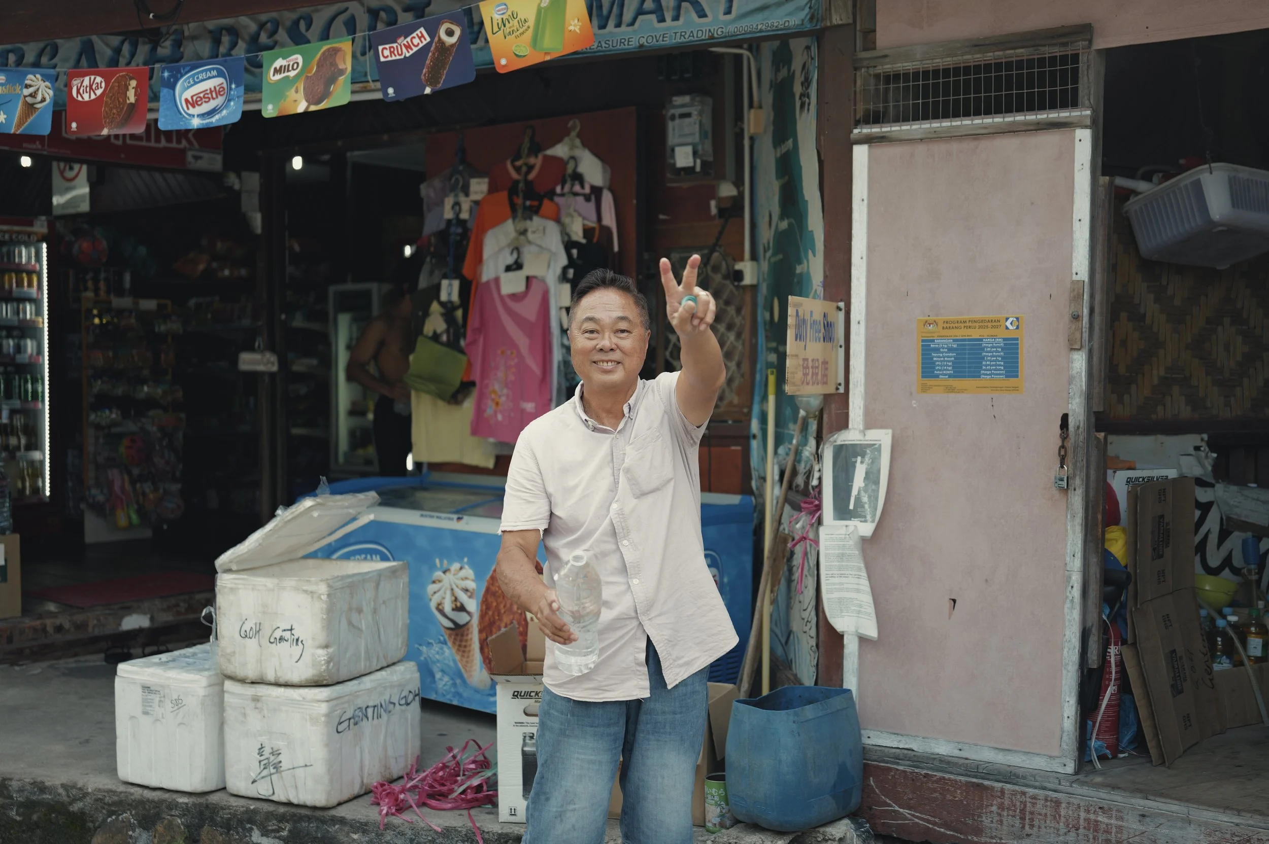 A smiling man in a white shirt and jeans standing outside a small store, pointing at the camera. The store has colorful ice cream advertisements hanging above, with several large white boxes and containers in front. The storefront is slightly open, r