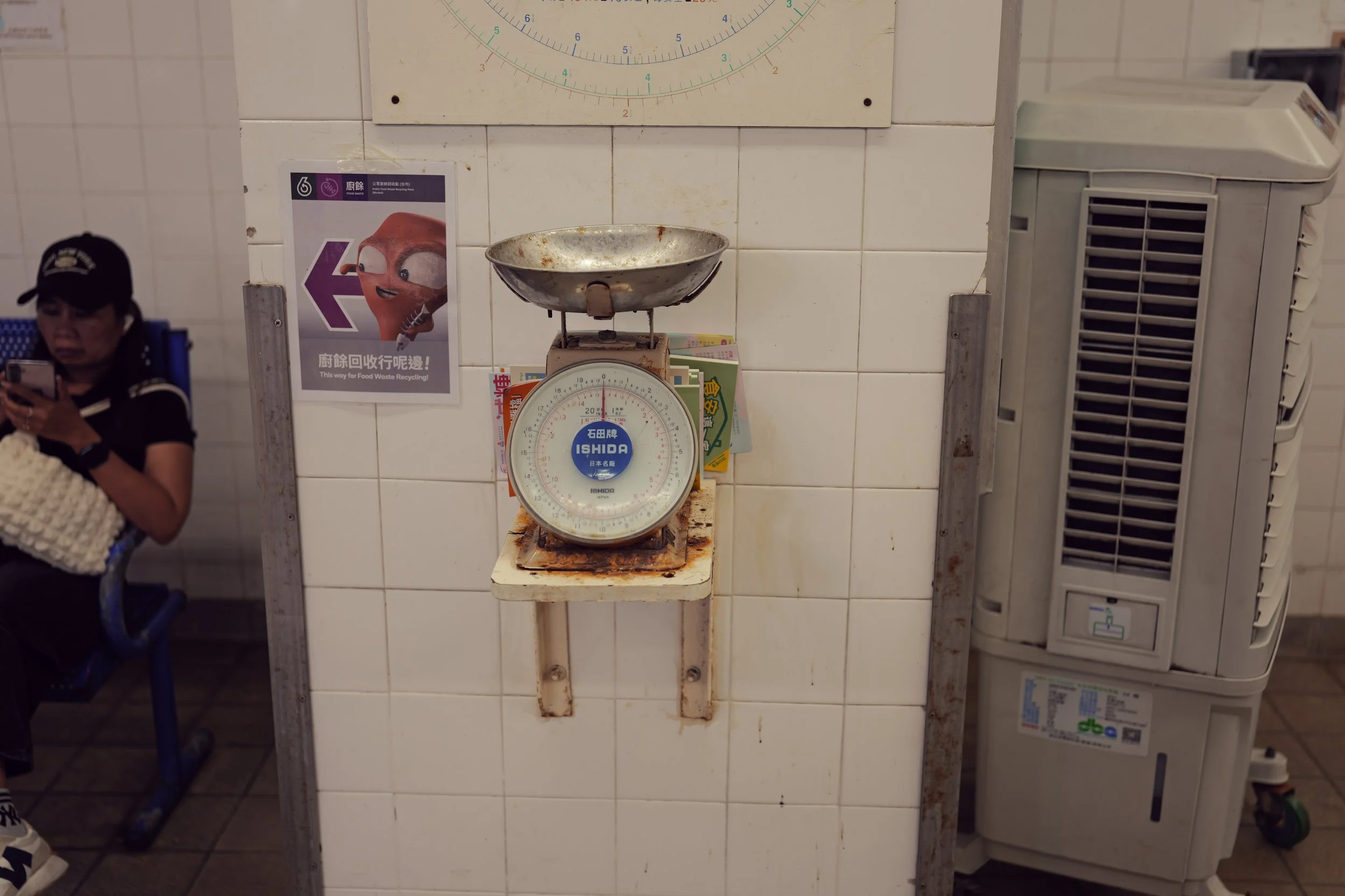 A vintage weighing scale with a round dial and a metal bowl on top, placed on a white tiled wall in a food establishment. An air conditioning unit is visible on the right side, and a person is sitting on the left side, looking at their phone.