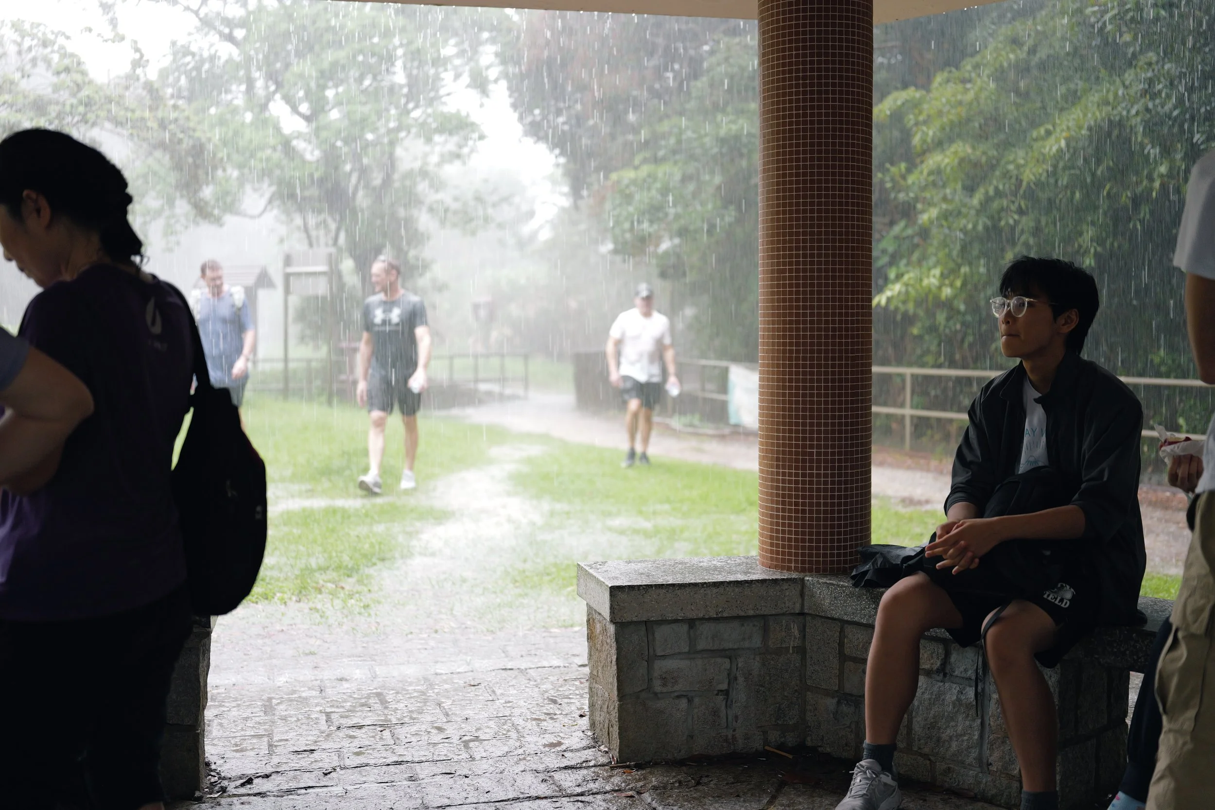 People sitting under a shelter watching rain outside, with some walking in the rain in the background.