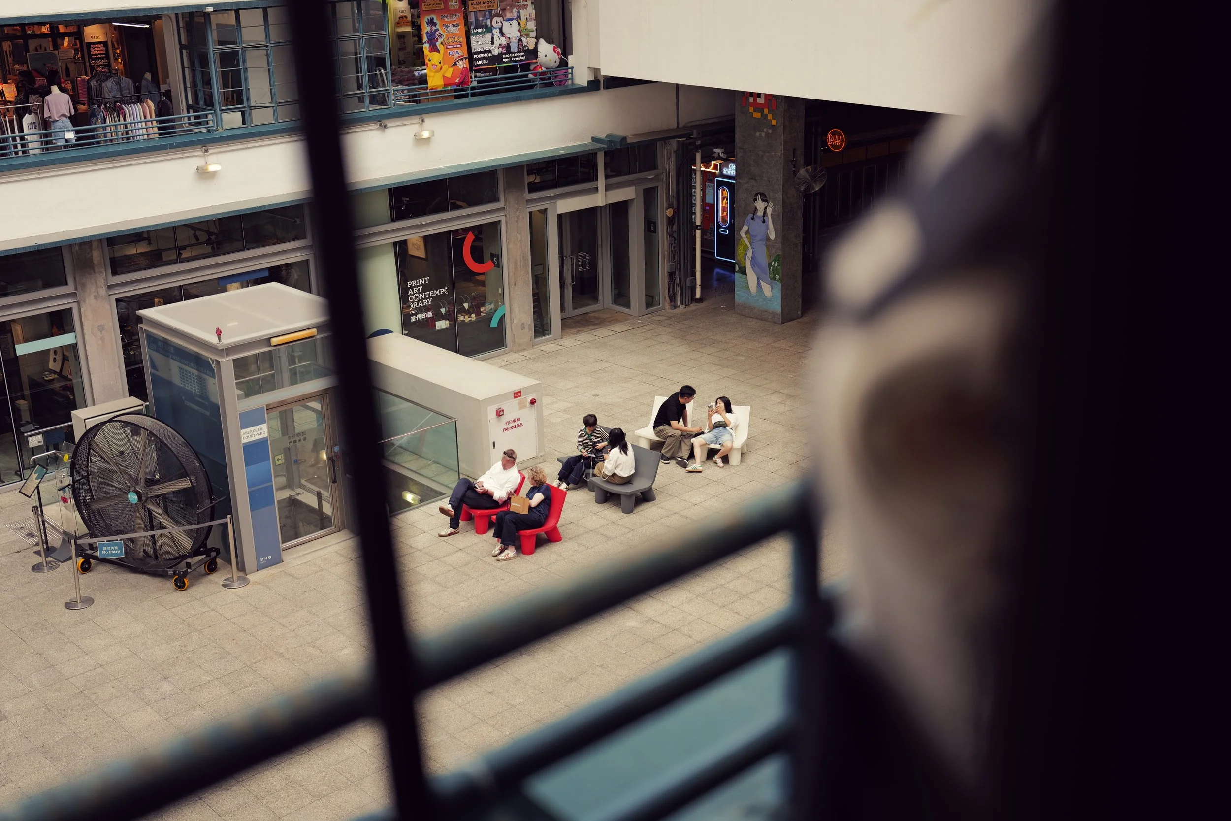 People sitting on benches in an indoor shopping mall courtyard. Some are engaged in conversation, others are using their phones. The mall has storefronts with display windows on the upper level and various colorful signage and figures.