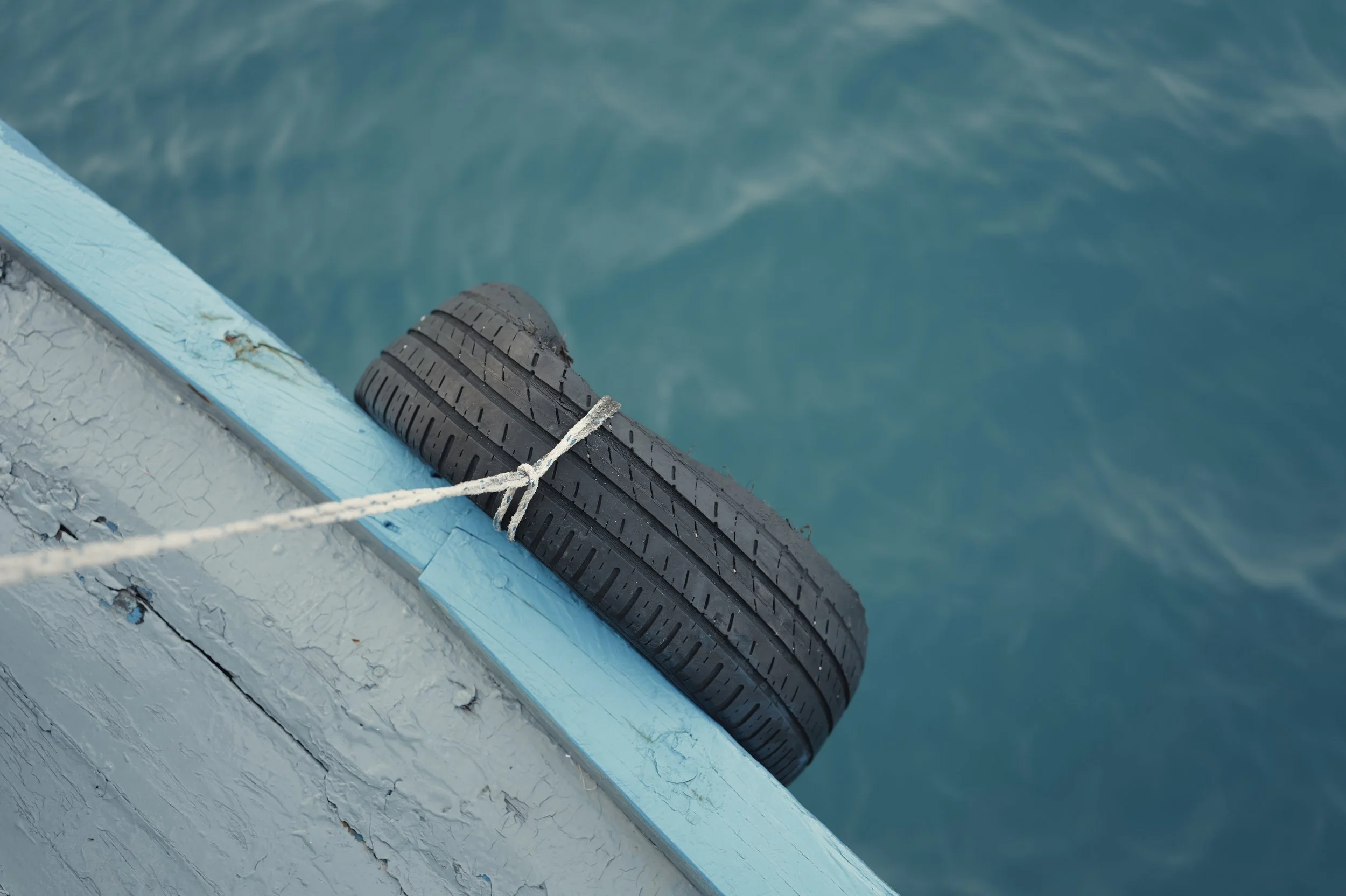 Close-up of a black rubber boat bumper tied with a white rope, attached to the edge of a weathered, light blue dock over turquoise water.