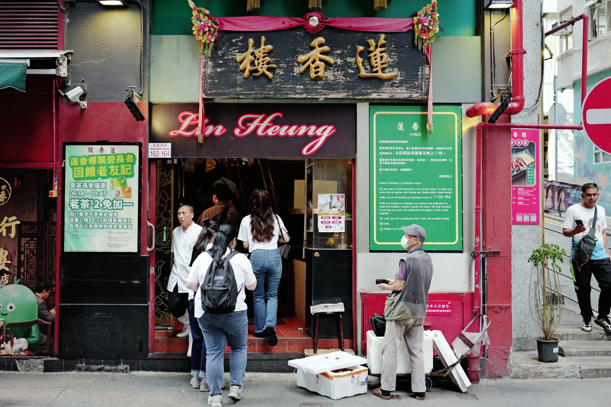 Street scene with a restaurant sign in Chinese characters, people waiting outside, and various colorful signs and decorations.