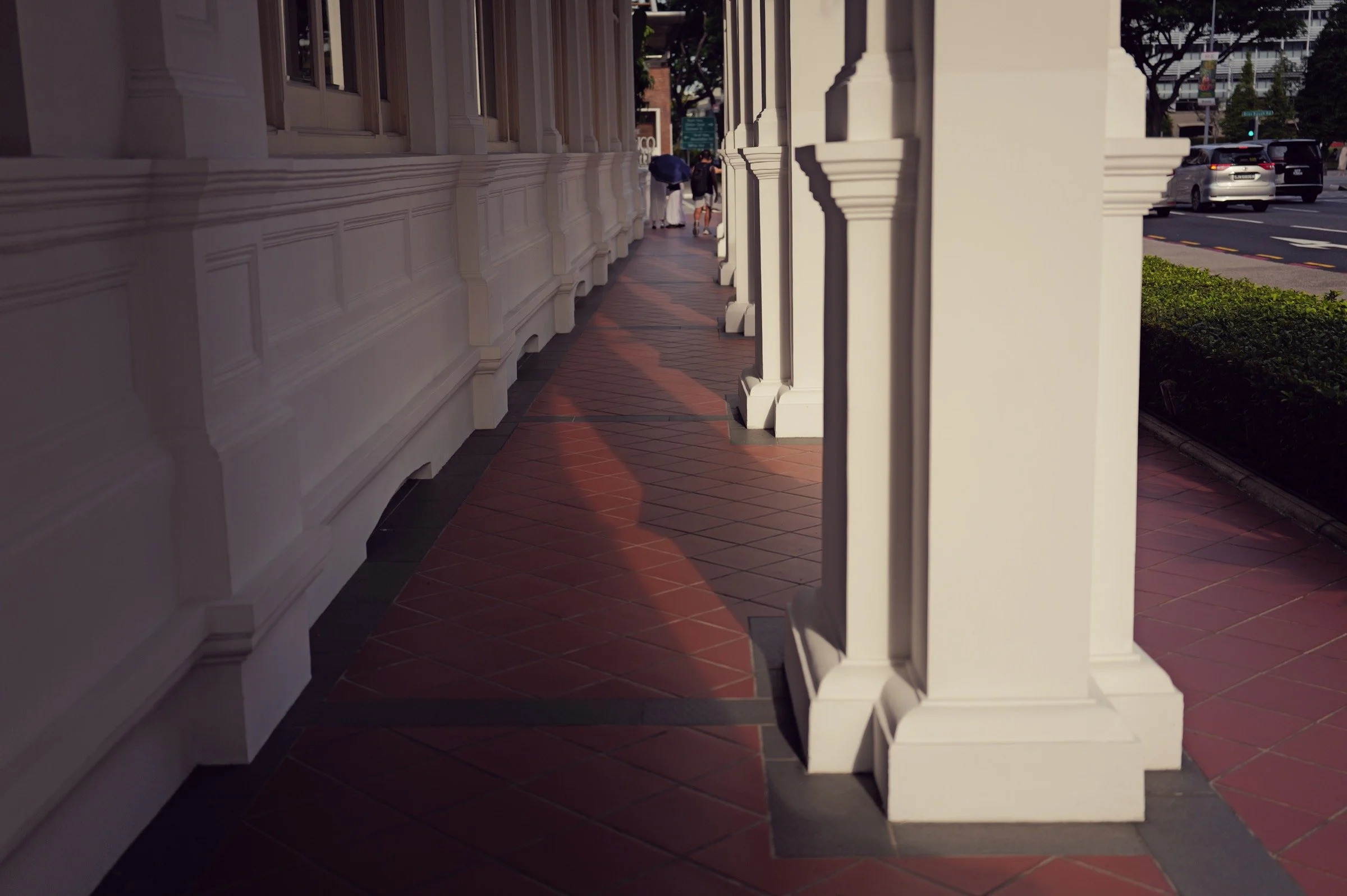 White columns cast long shadows across red and dark tiled flooring along a sheltered walkway.