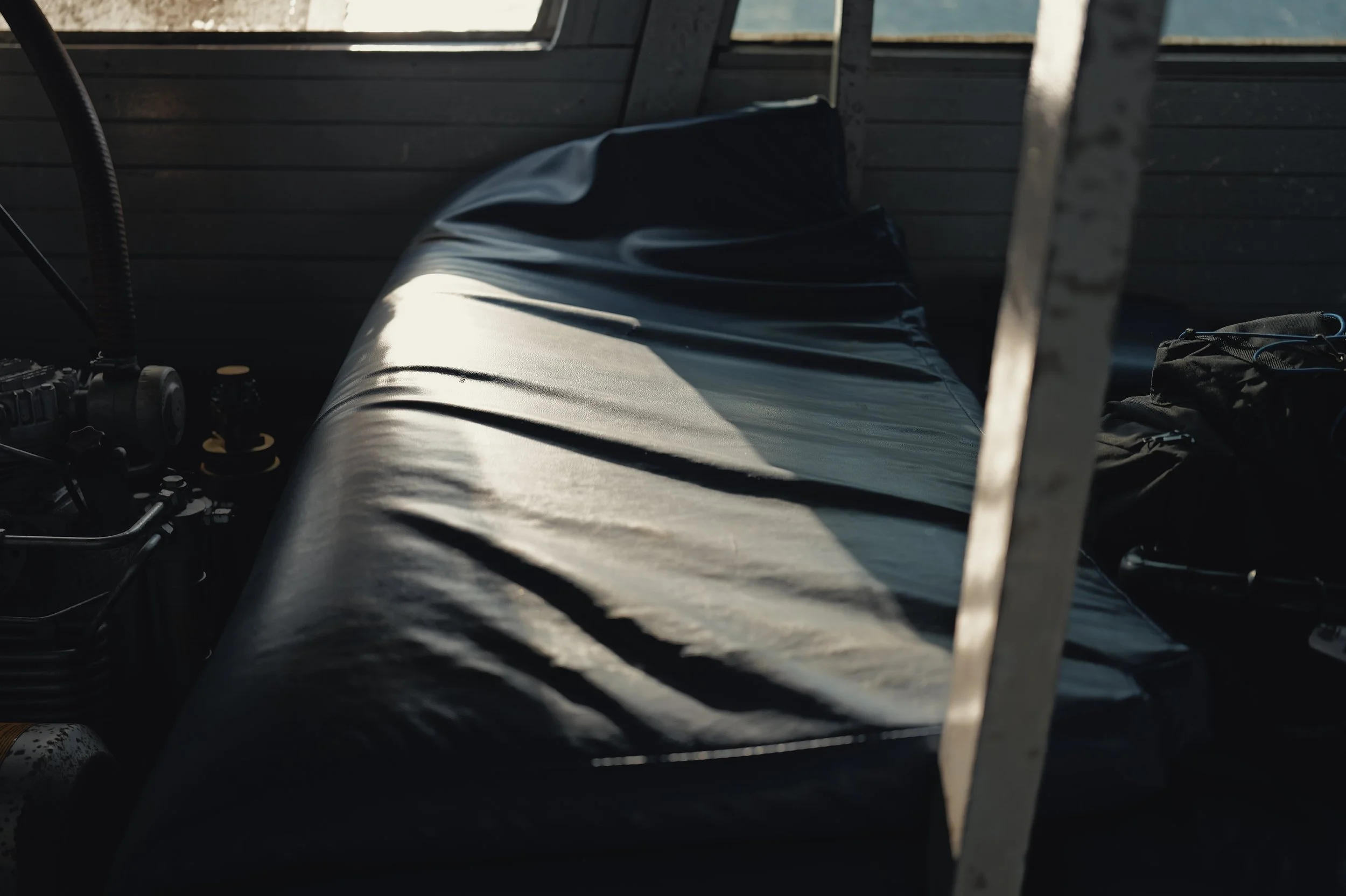 Close-up of a motorcycle with a black seat and parts, inside a dimly lit structure with wooden walls and a window, sunlight shining through.