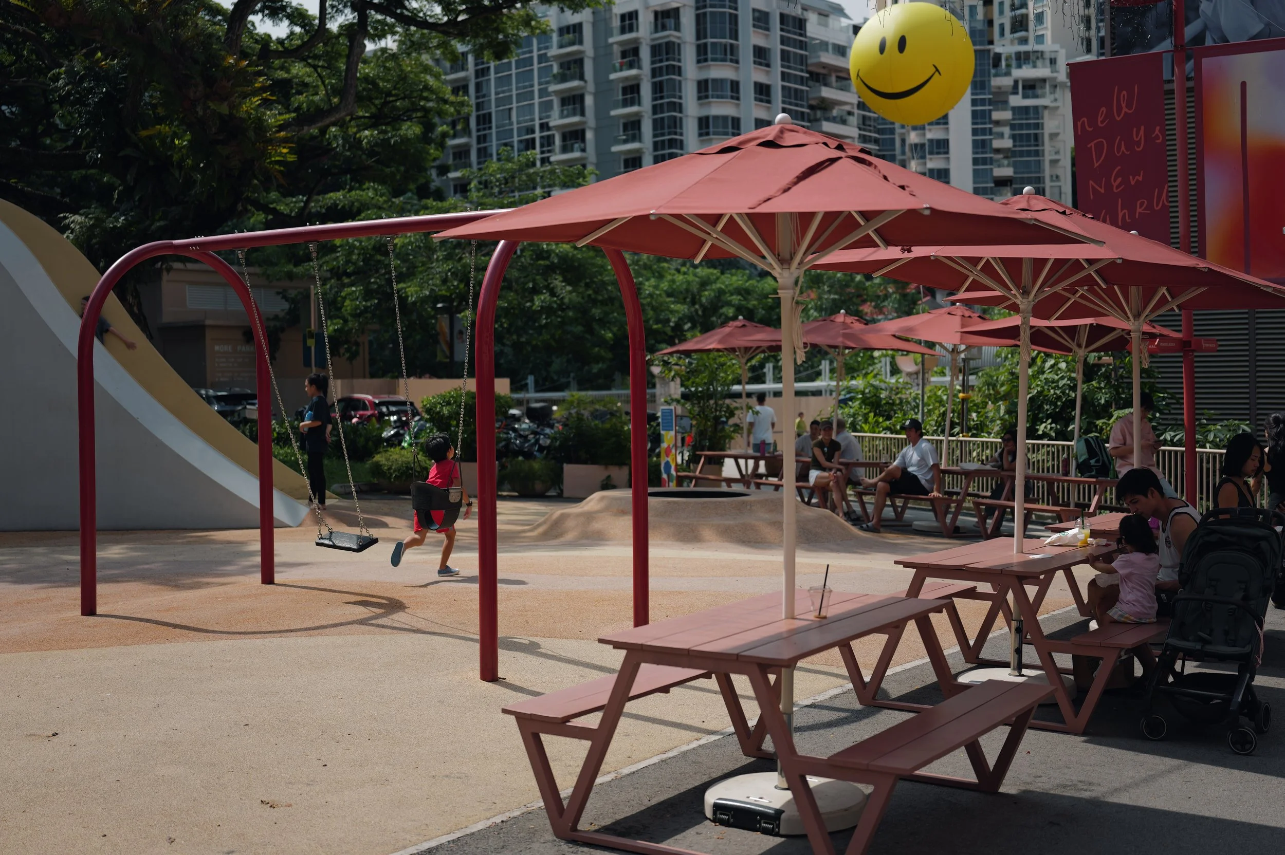 A playground in an urban park with pink umbrellas, a swing set, and children playing in the sun. People are sitting at picnic tables, and high-rise buildings are visible in the background. A large yellow smiley face balloon is floating in the air.