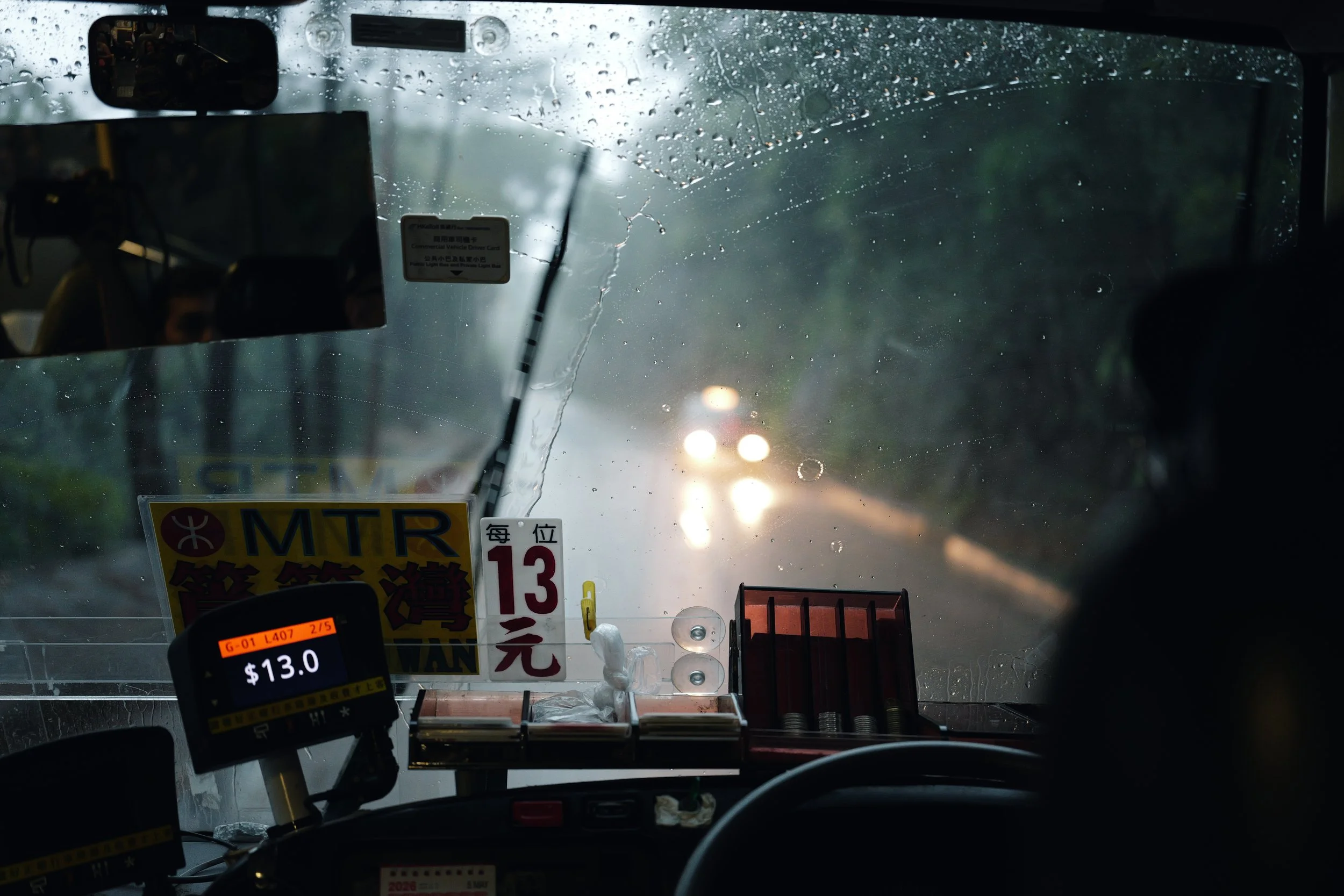 Inside a bus or taxi, rain on the windshield with traffic lights and blurred cars visible outside, and various cards and signs displayed inside the vehicle.