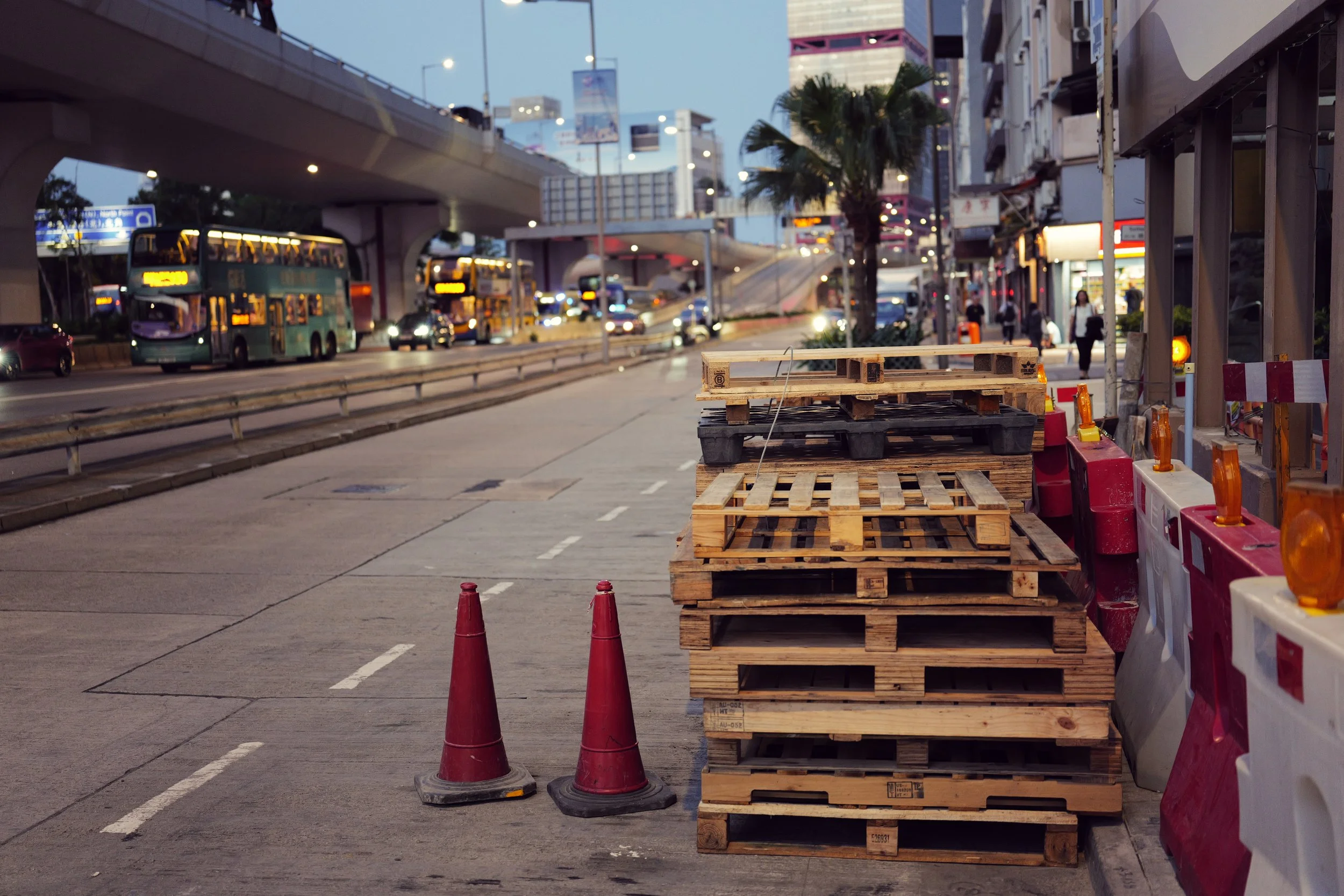Street scene with stacked wooden pallets, traffic cones, construction barriers, and a cityscape in the background with buses and high-rise buildings.