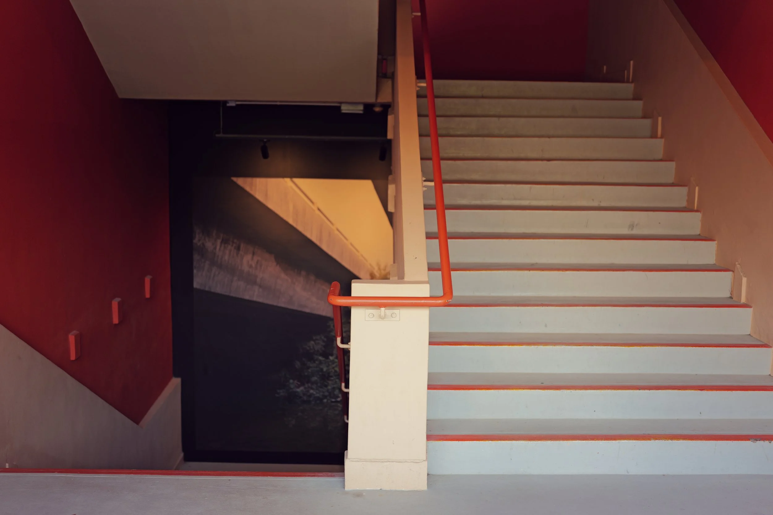 Interior view of a minimalist staircase with white steps, red handrails, a small corridor beneath the staircase, and a red wall on the left side.