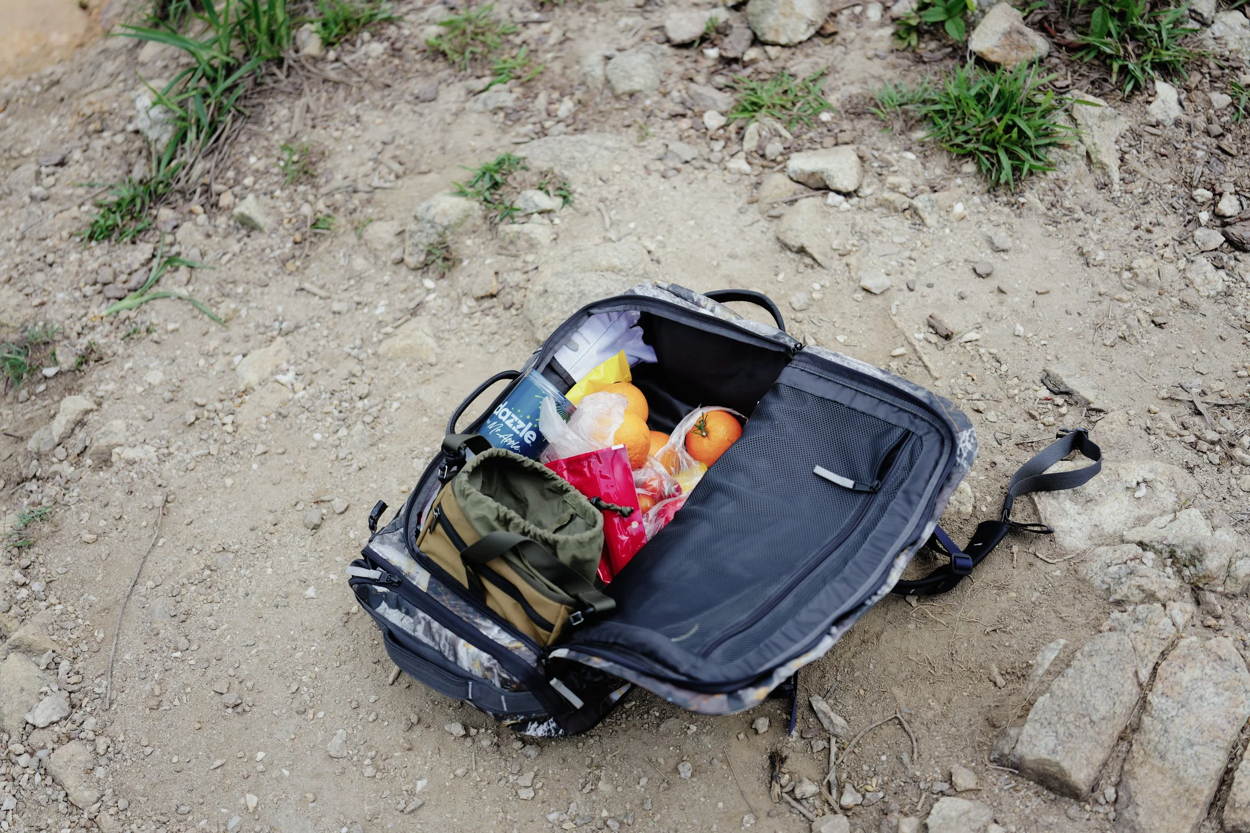 Open backpack on dirt ground with various food items including oranges and snacks, surrounded by small plants and rocks.