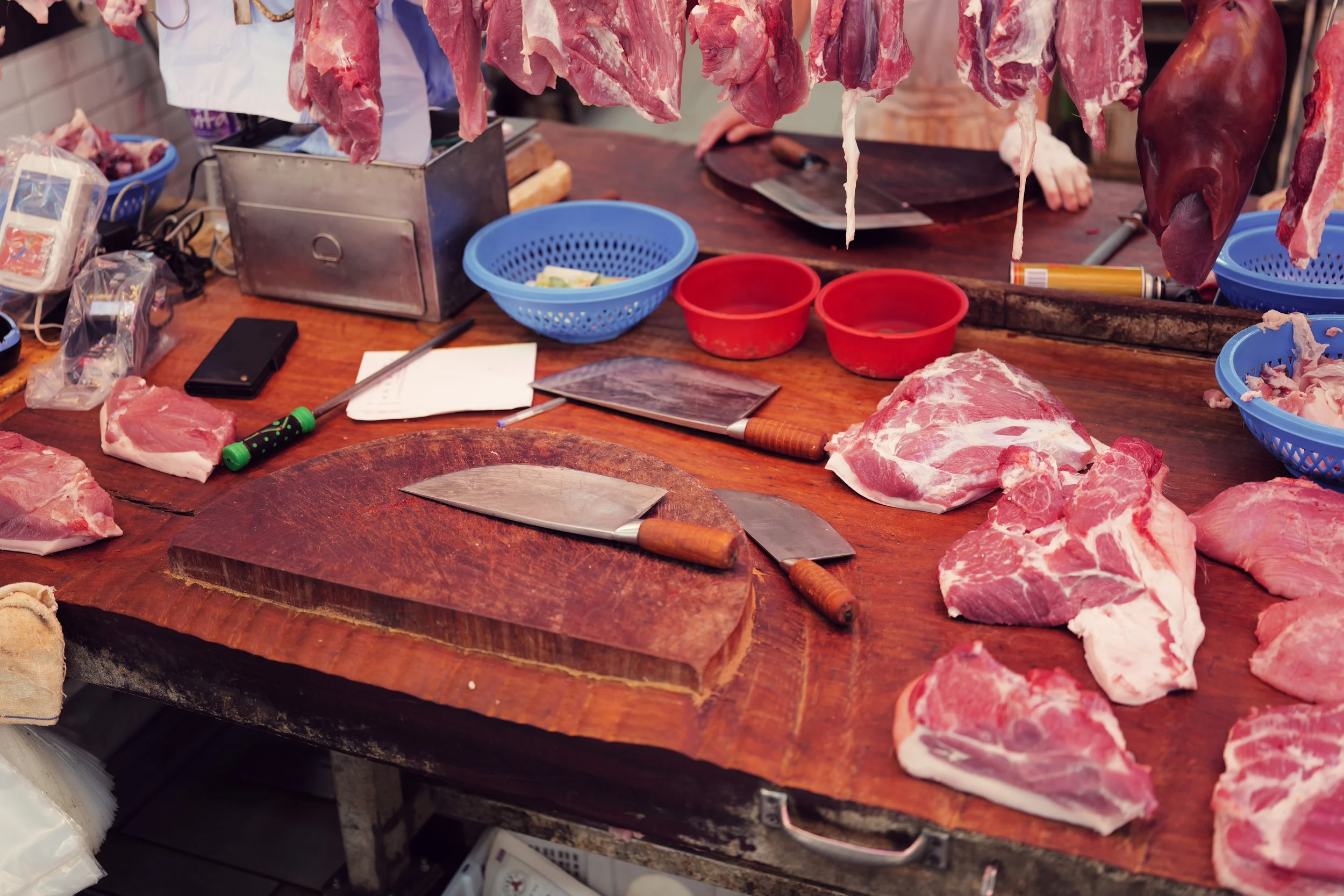 Various cuts of raw meat are displayed on a wooden table, with some hanging and others placed on the surface. There are knives, plastic baskets, and containers nearby, indicating a butcher shop or meat market setting.