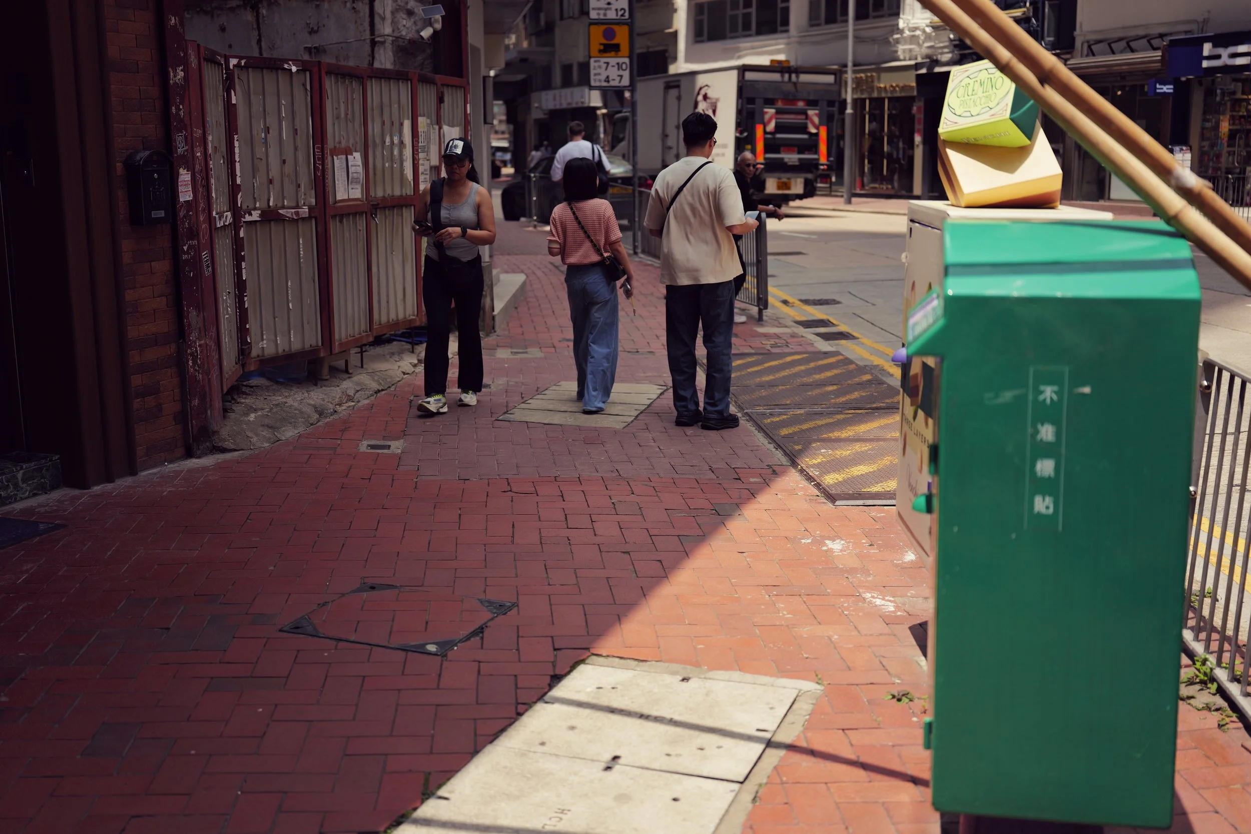 A city street scene with pedestrians walking on a brick sidewalk, a truck in the background, and colorful storefronts. There are pedestrians in casual clothing, some standing and others walking, with a focus on the brick sidewalk and urban environmen