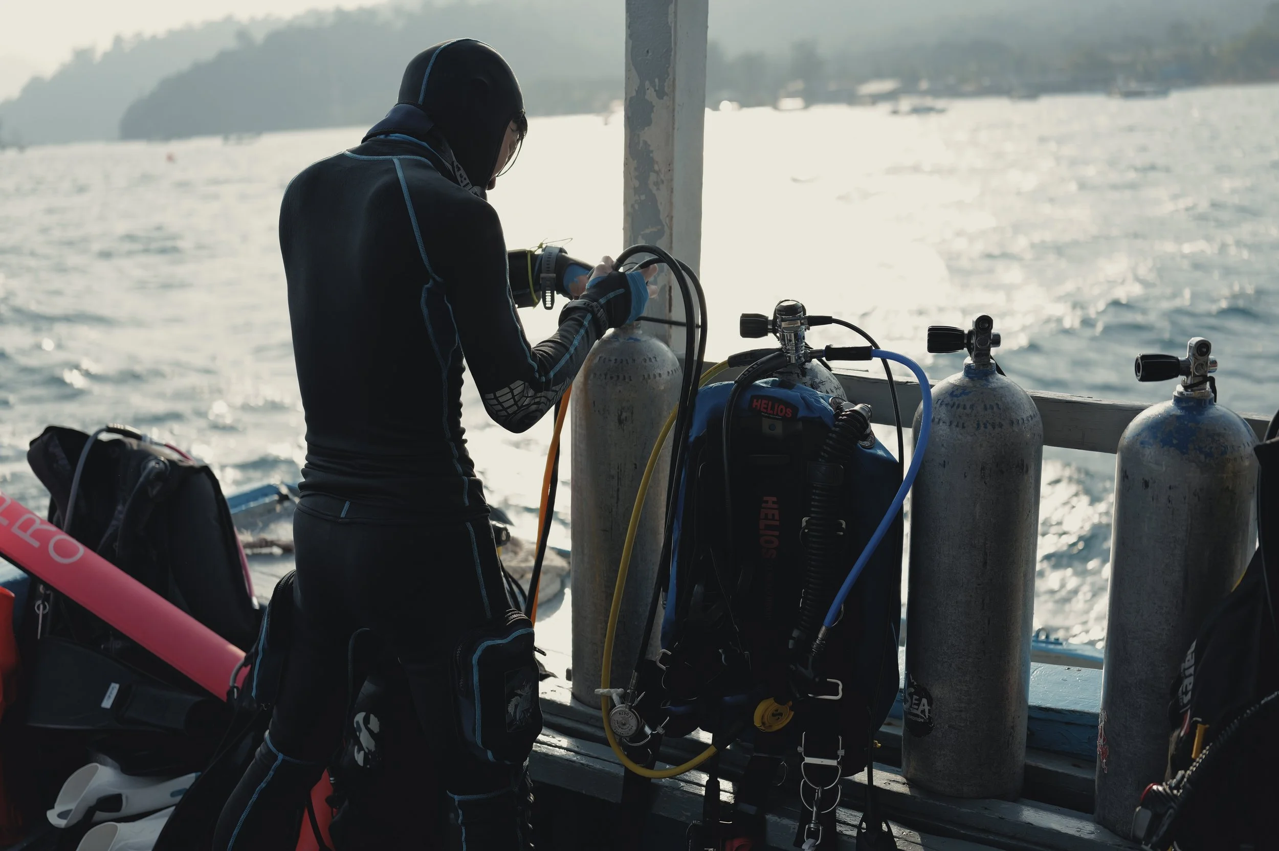 A person in a wetsuit and hood preparing scuba diving equipment on a boat near a body of water with a forested shoreline in the background.