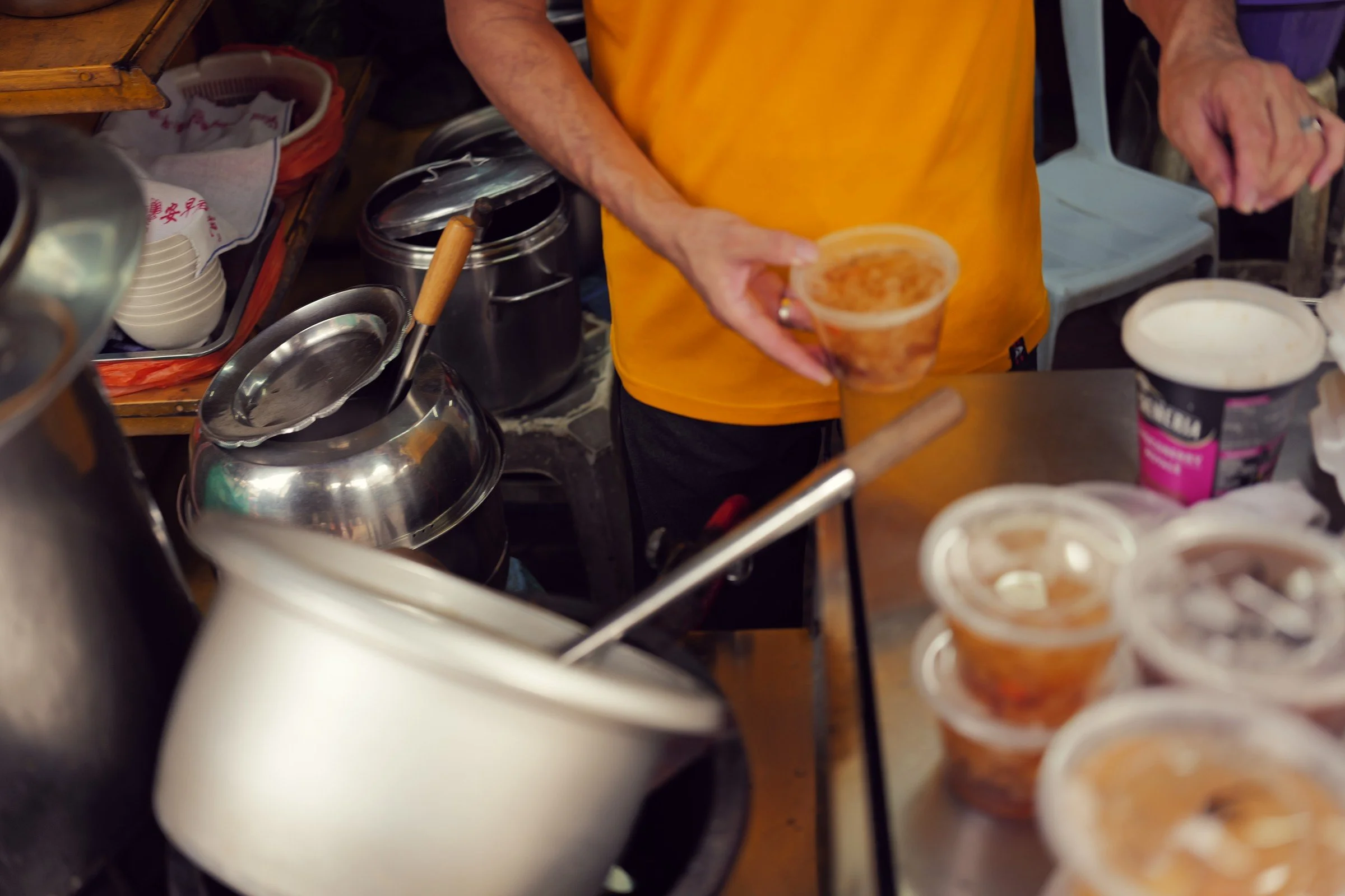 Hands prepare a drink among metal kettles and takeaway cups.