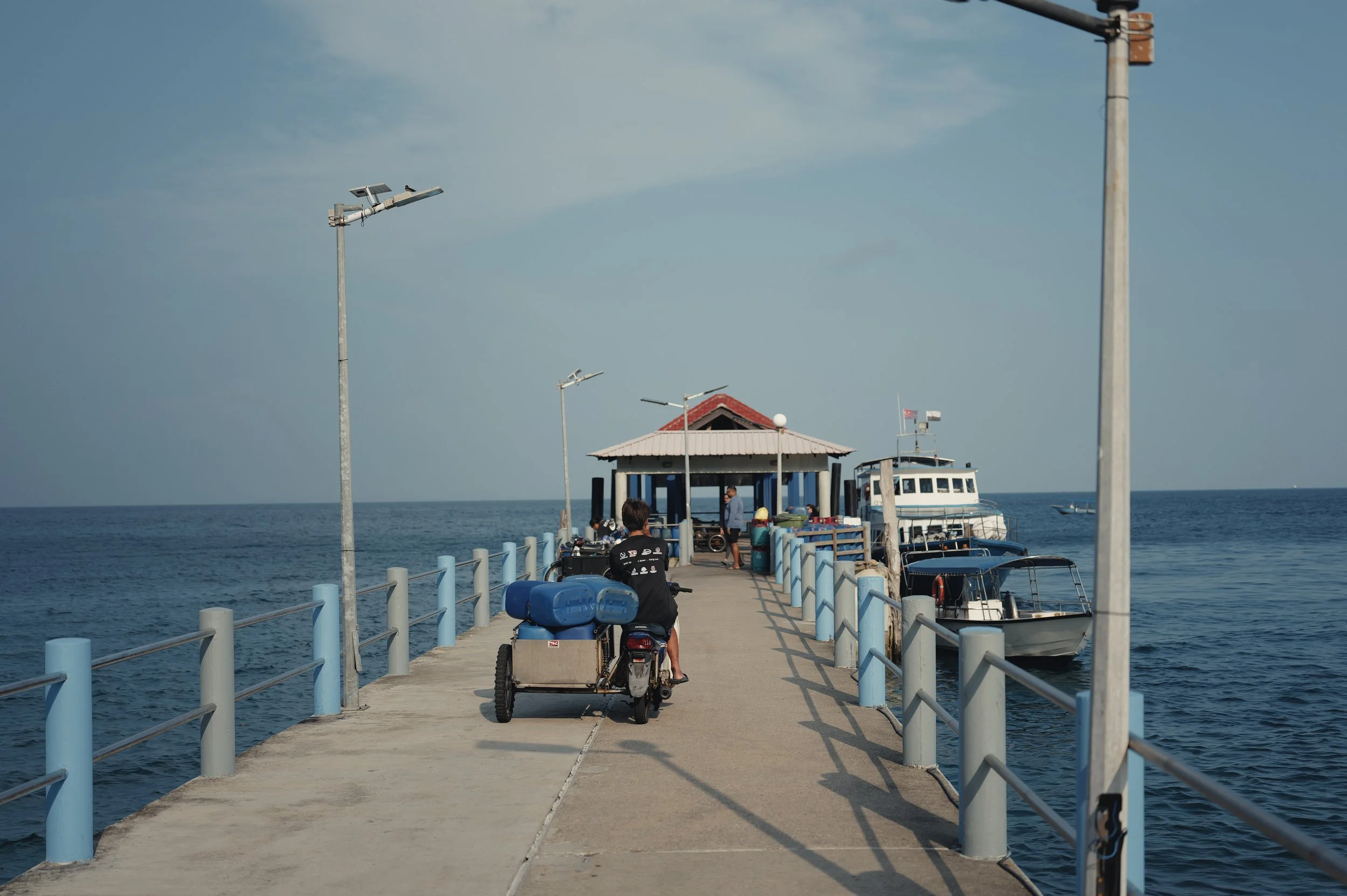 A pier extending into the ocean with boats docked on the right side, a small shaded area at the end, and a person riding an ATV on the pathway.