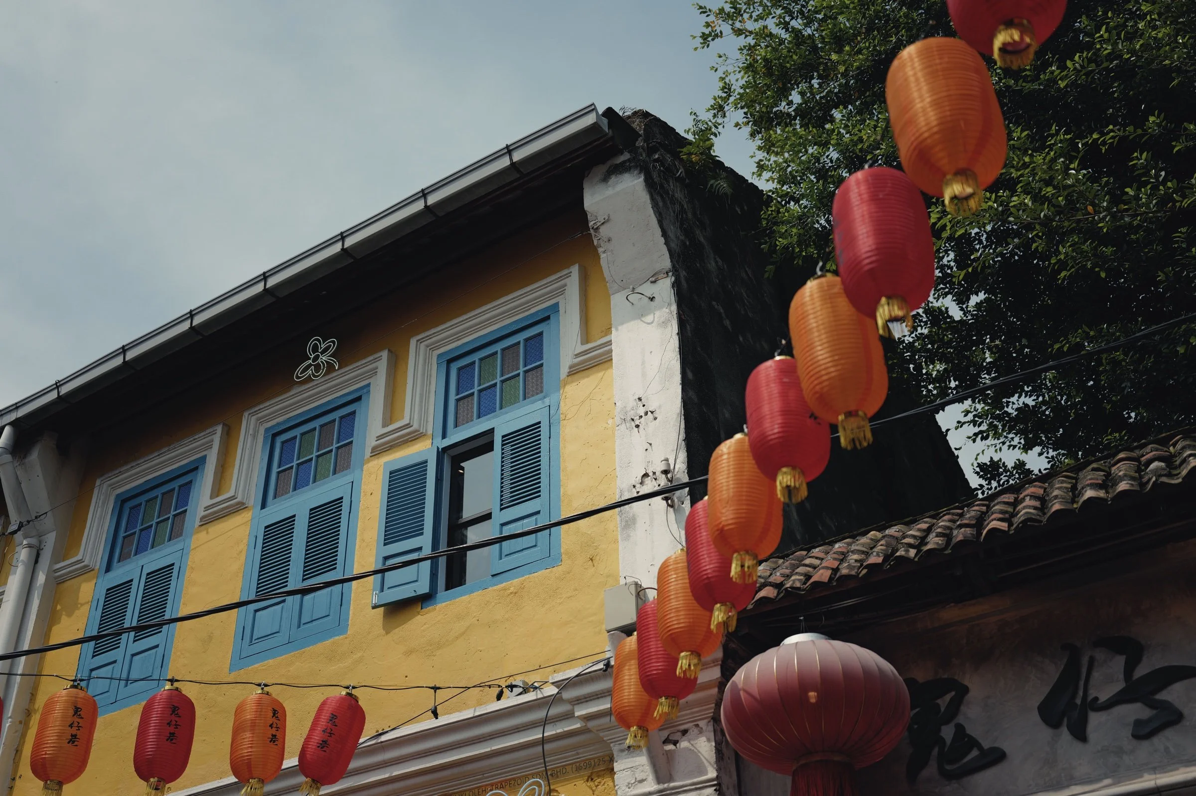 Red lanterns cross a yellow shophouse facade with blue shutters.