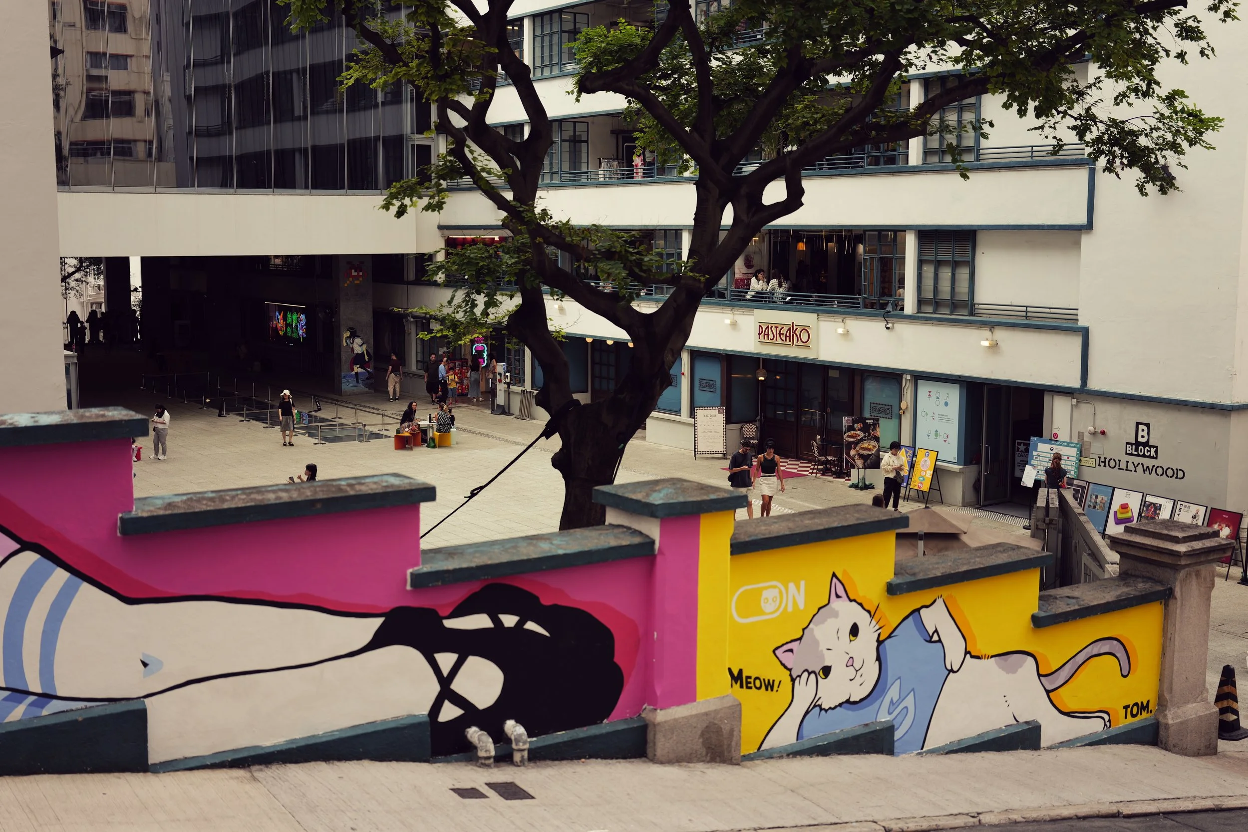 View of an outdoor urban plaza with people walking, sitting, and socializing, surrounded by a modern building with multiple levels and glass windows. A large tree is in the center, and there are colorful decorative elements on the wall in the foregro