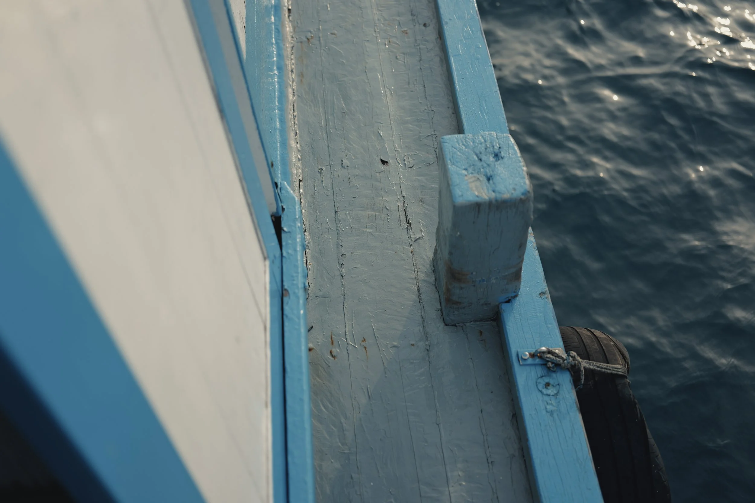 Close-up of the side of a boat with chipped blue and white paint, showing part of the boat's edge and a tire attached as a bumper, with water in the background.