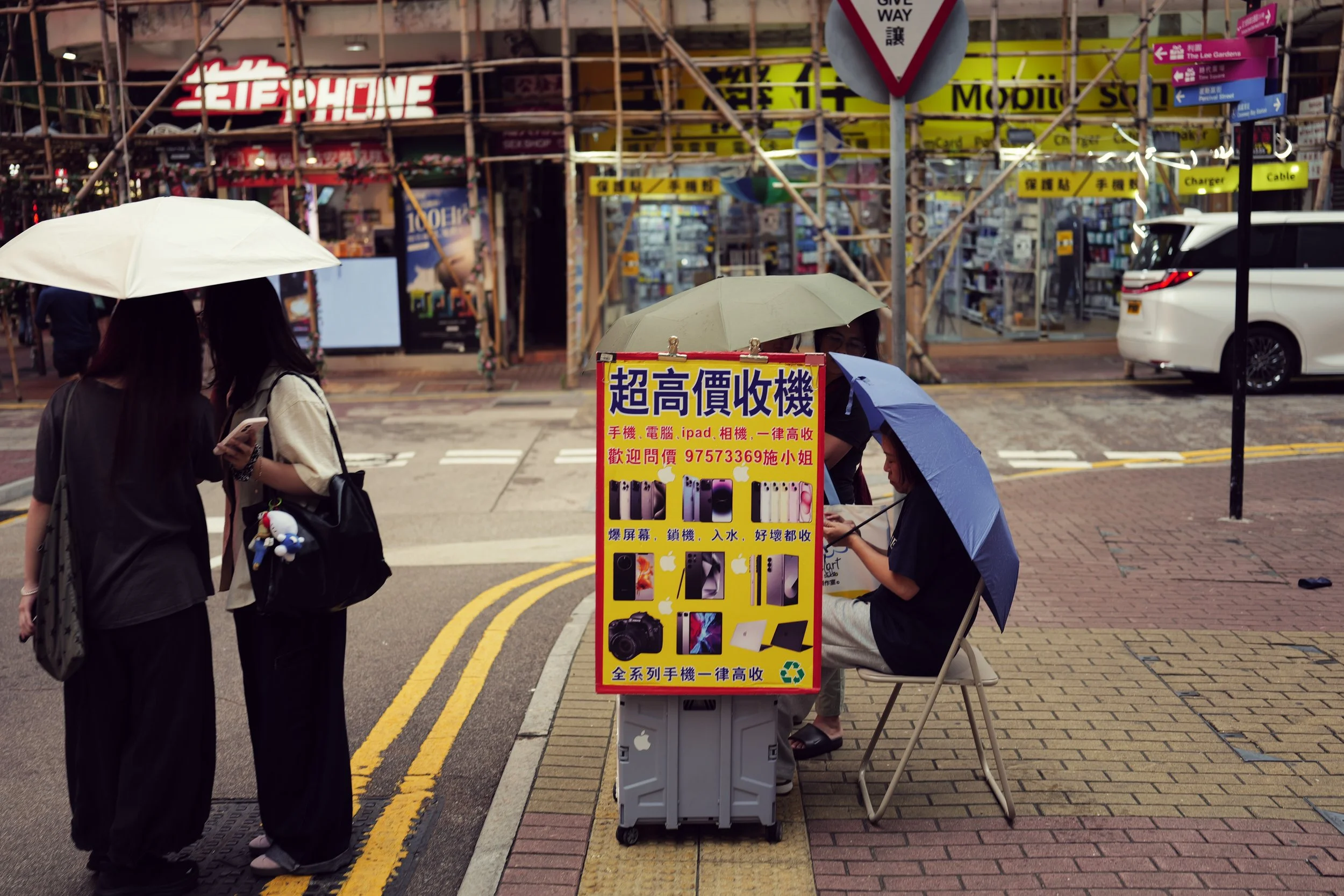 Street corner with a mobile phone accessory stall, two women holding umbrellas, and a building with scaffolding. One woman is sitting behind the stall, which has a yellow sign advertising phone and electronic device recycling in Chinese. A white car 