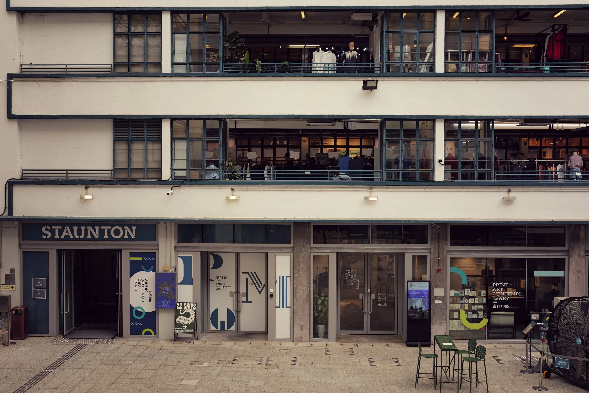 Front of a multi-level commercial building with glass storefronts, displaying clothing and art-related signage, an outdoor seating area, and a tiled sidewalk in front.