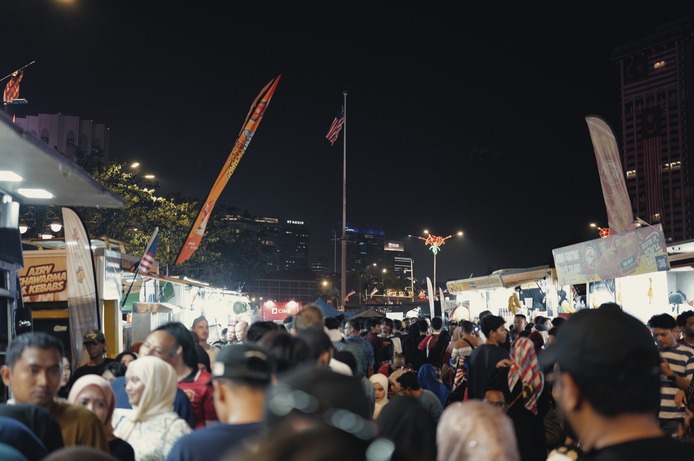A crowd fills a brightly lit night market beneath tall flags and the dark sky.