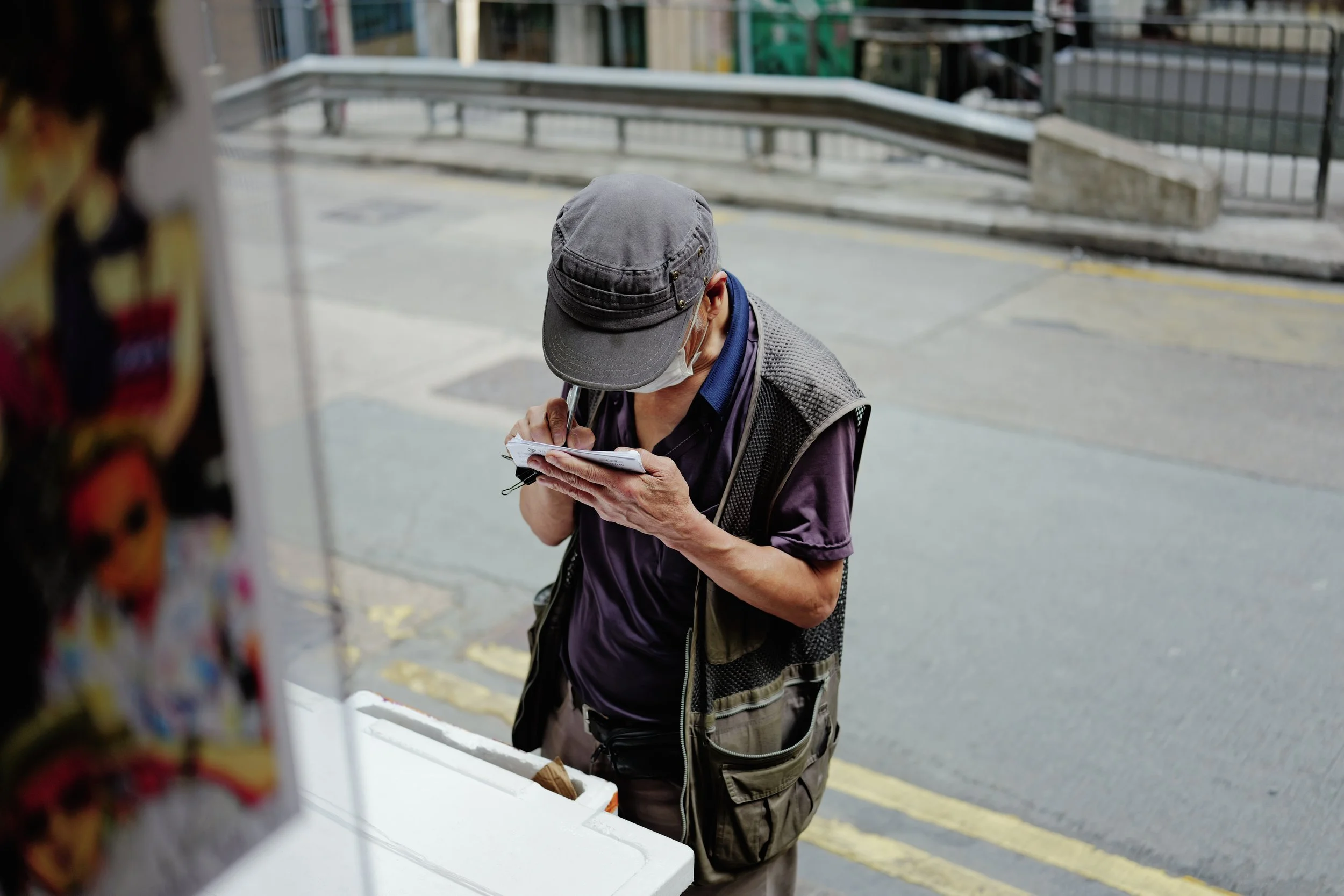 Older man wearing a gray cap and face mask reviewing papers on a clipboard outside in an urban setting.